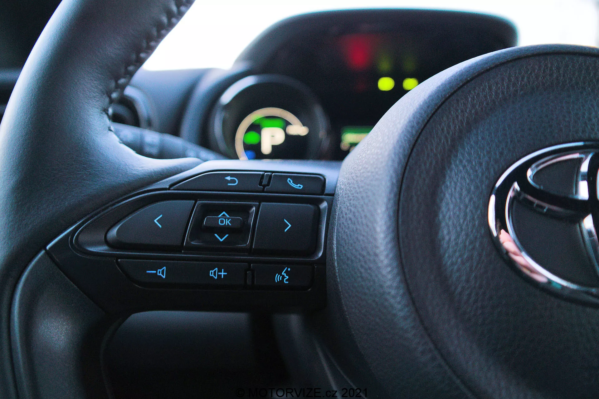 Vista interior en primer plano de un Toyota Yaris 2020 desde la perspectiva del asiento del conductor, destacando el volante con controles multifunción y un cuadro de instrumentos en el fondo. El volante domina los dos tercios inferiores de la imagen, con flechas, comando de voz, volumen y botones del sistema de entretenimiento visibles. El cuadro de instrumentos parcialmente visible está iluminado con una pantalla digital verde y está desenfocado debido a la poca profundidad de campo. La luz natural ilumina el interior, enfatizando la comodidad y una interfaz fácil de usar.