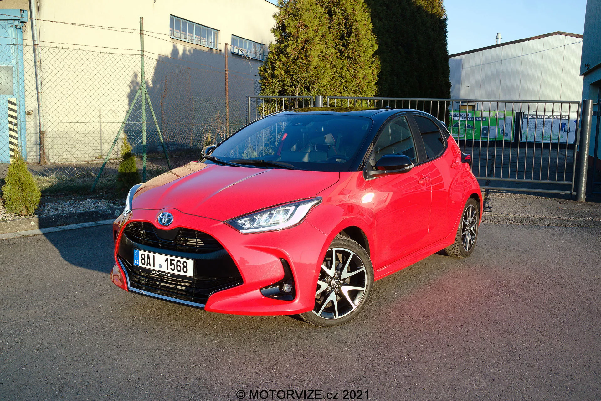 Imagen de un hatchback Toyota Yaris 2020 rojo brillante estacionado en ángulo, mostrando tanto el perfil delantero como el lateral. El diseño de los faros es afilado y angular, la parrilla tiene un patrón de panal con un emblema centralizado, y el parachoques delantero tiene contornos visibles con luces antiniebla. El perfil lateral revela llantas de aleación de dos tonos, manijas y espejos del color del cuerpo con líneas y contornos nítidos a lo largo del costado. La pintura brillante sugiere un estado nuevo o bien mantenido. El fondo incluye un cielo claro, estructuras industriales y sombras suaves, indicando un entorno al aire libre, probablemente un área de estacionamiento cerca de una instalación comercial o industrial.