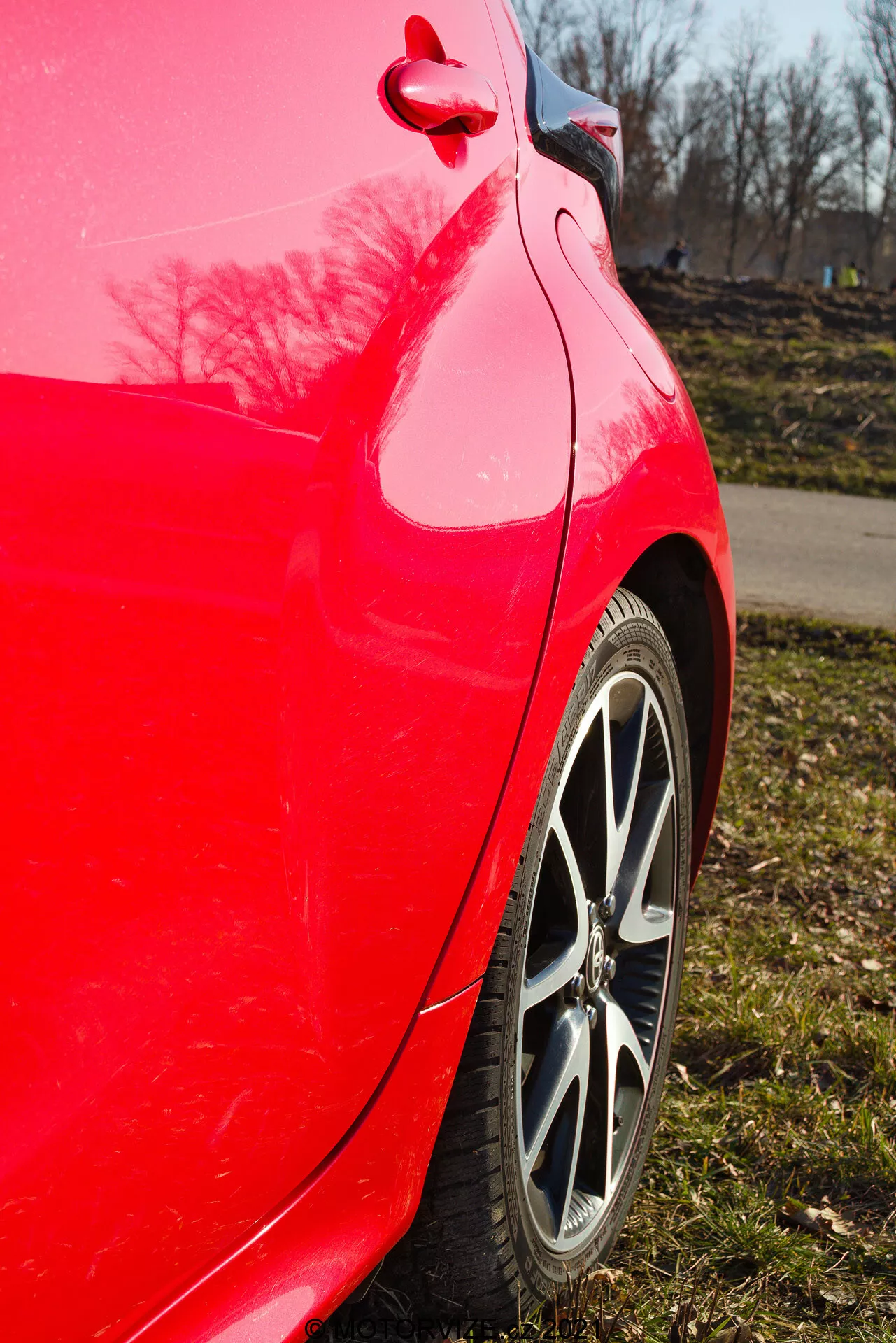 Vista lateral de cerca del panel trasero del Toyota Yaris 2020 rojo tomada en un ángulo ligeramente ascendente resaltando el acabado rojo brillante, la llanta de aleación de múltiples radios, el patrón de la banda de rodadura del neumático, la curva limpia del panel trasero y el parachoques, el reflejo brillante de la pintura con árboles y un cielo azul claro, y la tapa de combustible y el detalle de la moldura visibles.