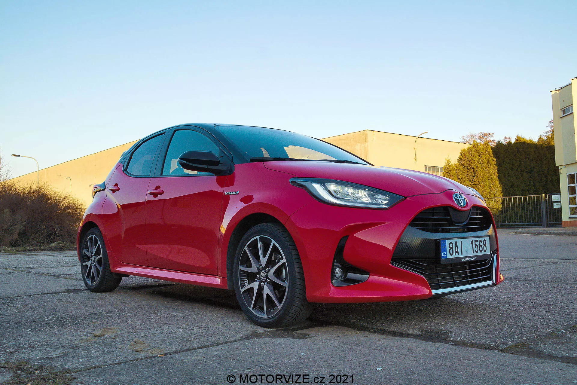 Vista de tres cuartos delanteros a baja altura de un Toyota Yaris hatchback 2020 de color rojo vibrante que muestra faros LED o HID angulares y nítidos, una parrilla prominente con emblema central, ruedas de aleación con múltiples radios, señales de giro integradas en los espejos laterales, superficies reflectantes limpias y un posible sistema de frenos orientado al rendimiento. Capturado en un área de estacionamiento industrial con iluminación cálida y marca de agua motorvize.cz 2021.