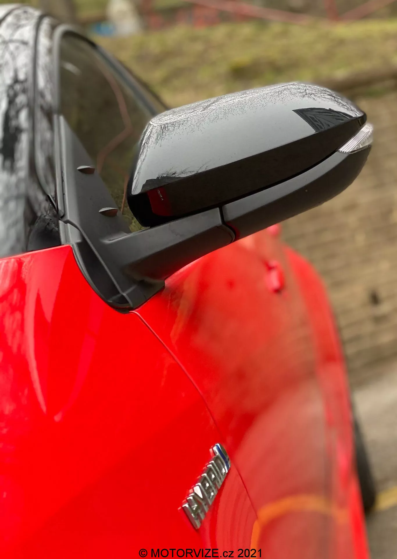 Primer plano del espejo lateral desplegado de un Toyota Yaris 2020, con un diseño aerodinámico negro que contrasta con la carrocería roja y brillante del coche. El espejo destaca posibles características tecnológicas como ajuste eléctrico, calefacción y señales de giro integradas, aunque no se confirman características específicas. Las gotas de agua sugieren lluvia reciente o lavado de coche, mientras que un fondo desenfocado y poca profundidad de campo mantienen el enfoque en el espejo. Parte de una insignia en el guardabarros delantero es visible pero está desenfocada.