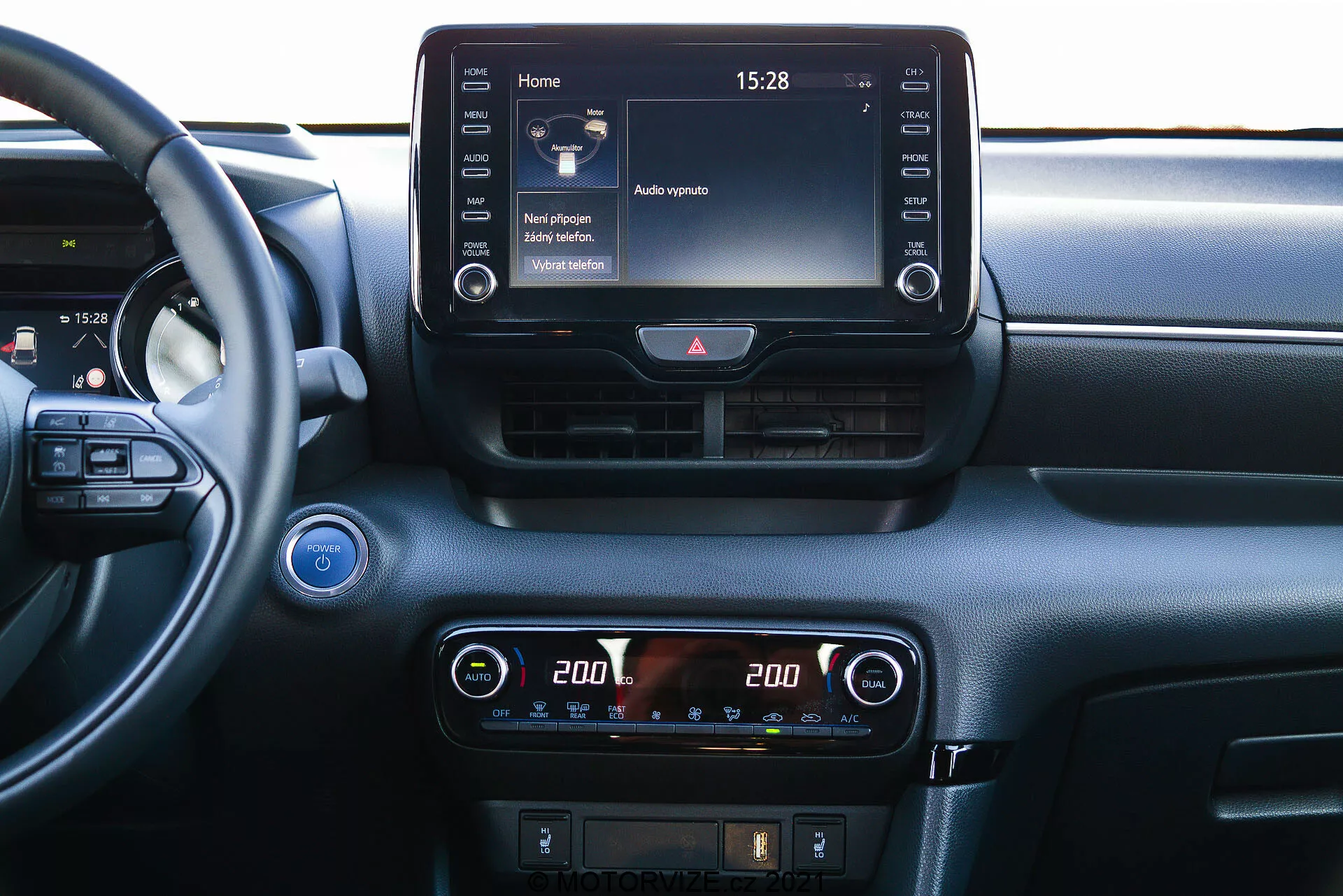 Vista de primer plano del tablero central de un Toyota Yaris 2020 desde una perspectiva interior. El volante es parcialmente visible a la izquierda, sugiriendo un ángulo de visión desde el asiento del pasajero o central. El tablero presenta una pantalla táctil prominente de infoentretenimiento en la parte superior de la consola central, mostrando una pantalla de inicio con varios iconos y la hora '15:28'. Debajo, un control de climatización digital con ajuste de temperatura de doble zona, cada lado configurado a '20.0' grados Celsius. Controles adicionales para el desempañamiento y posiblemente asistencia al conductor son visibles debajo de los controles de clima. Encima de la pantalla de infoentretenimiento, hay dos ventilaciones horizontales con un botón de luces de emergencia entre ellas. Un botón de encendido/apagado es parcialmente visible en el extremo izquierdo, indicando tecnología de ignición sin llave. Una rejilla de altavoz se encuentra en la esquina superior izquierda. El material del tablero cuenta con elementos lisos y texturizados, sugiriendo un enfoque en confort y tecnología.