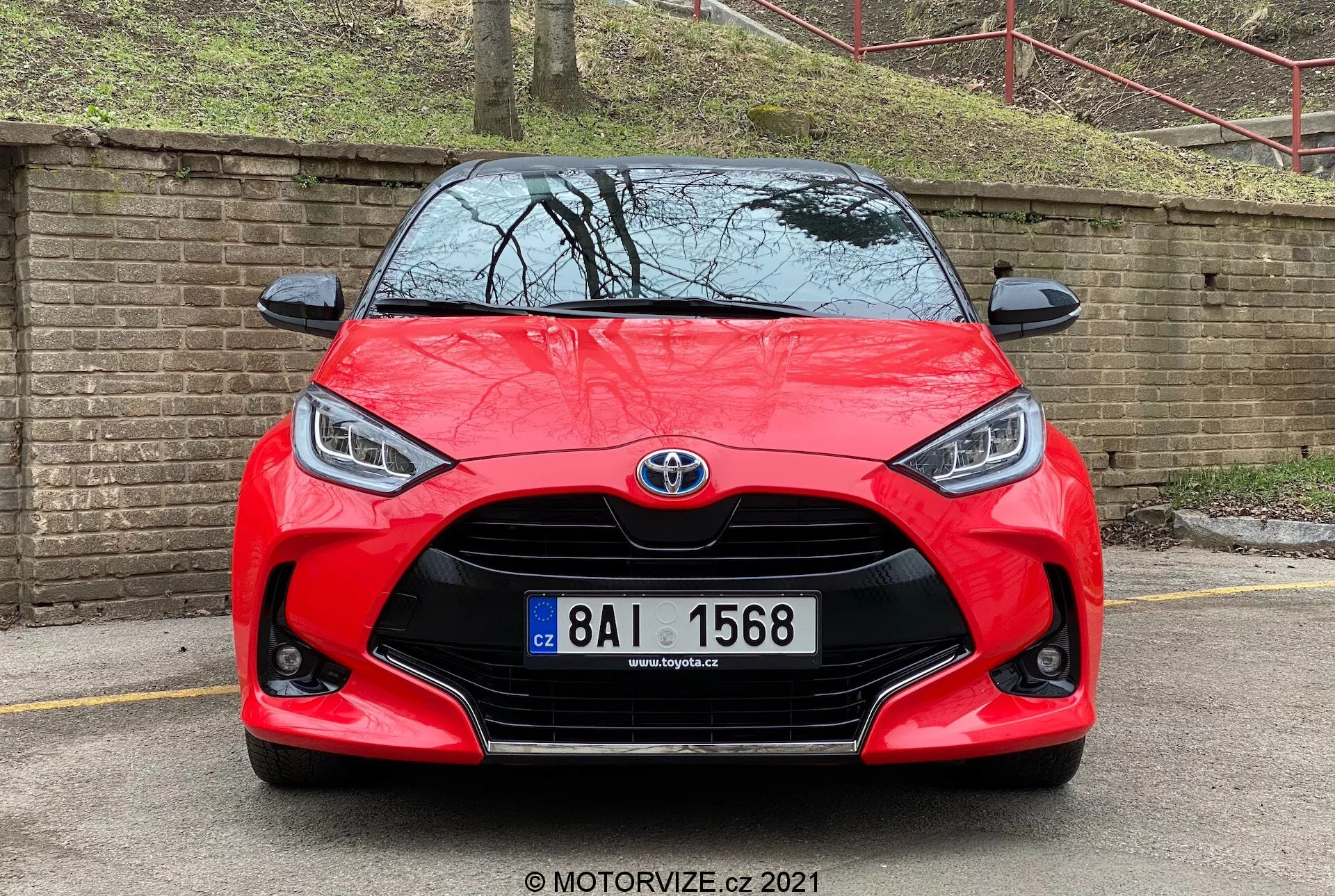 Vista frontal de un Toyota Yaris rojo 2020 que muestra su distintiva parrilla frontal con un prominente emblema de Toyota en el centro. La parrilla con diseño de rejilla está flanqueada por faros angulares elegantes que cuentan con luces diurnas LED. Debajo de la parrilla hay un conducto de aire visible rodeado por carcasas de luces antiniebla. El capó esculpido y el paragolpes delantero de integración perfecta contribuyen al diseño deportivo del coche. La placa de matrícula está montada en el centro, y la imagen tiene una marca de agua de MOTORVIZE.cz, fechada en 2021.
