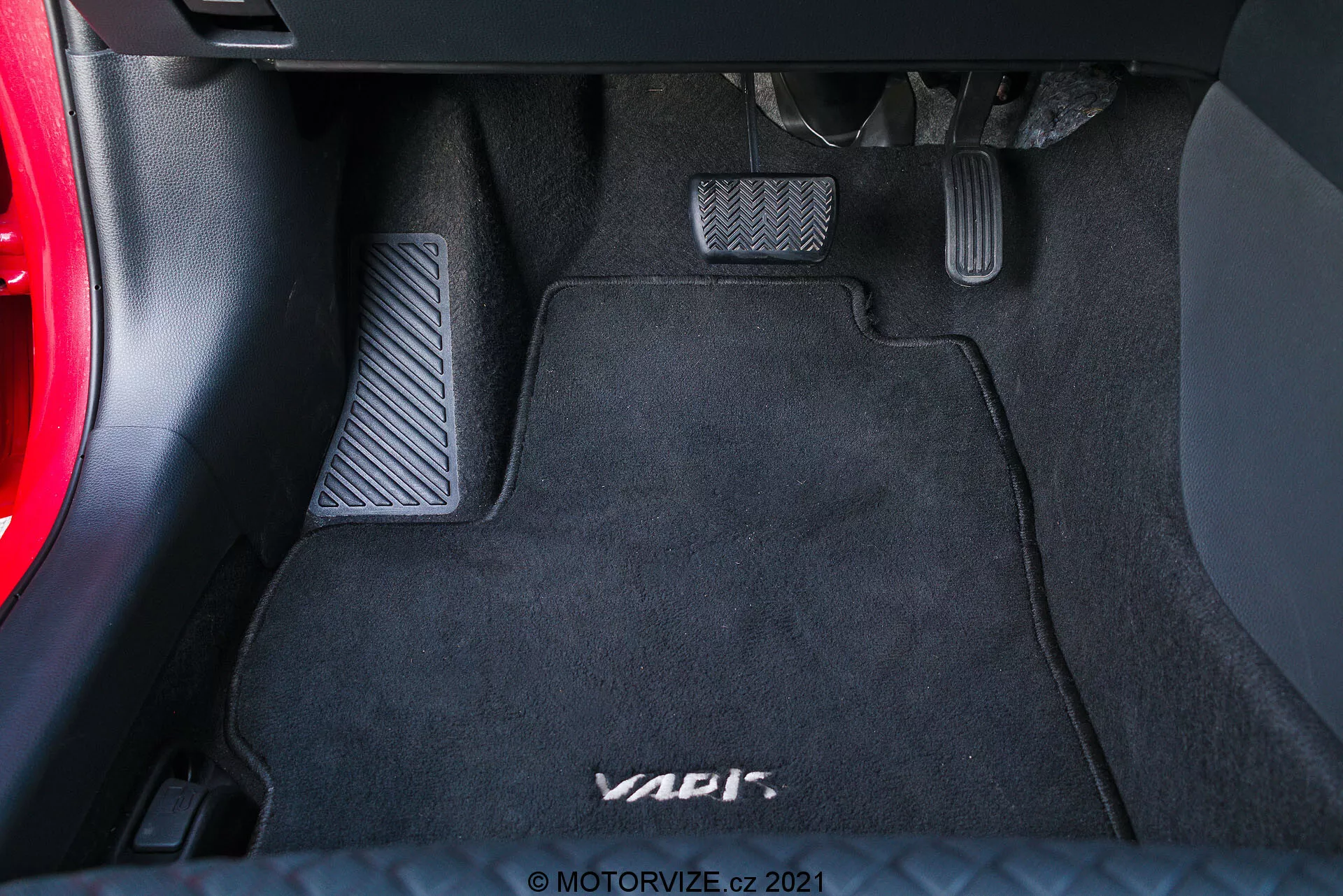 Vista de cerca del área del reposapiés del lado del conductor en un vehículo, destacando tres pedales con acabado metálico y insertos de goma para una configuración de transmisión manual en una alfombrilla de suelo negra con la marca 'VAG'. El pedal izquierdo es el embrague, el del medio es el freno y el de la derecha es el acelerador. La parte inferior del tablero y una sección de la puerta del coche son parcialmente visibles.