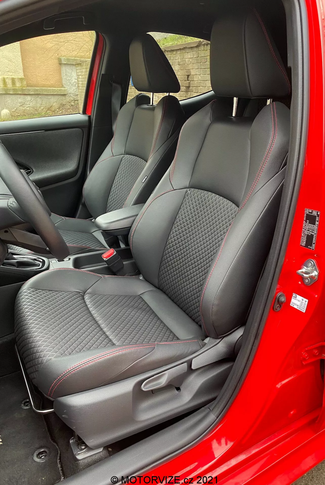 Interior del Toyota Yaris 2020 visto desde el lado del pasajero con la puerta abierta, destacando los asientos de tela texturizada premium con patrones de costura en forma de diamante y detalles en rojo, reposacabezas ajustables, controles de la consola central y panel de puerta con textura de fibra de carbono y controles eléctricos de las ventanas.