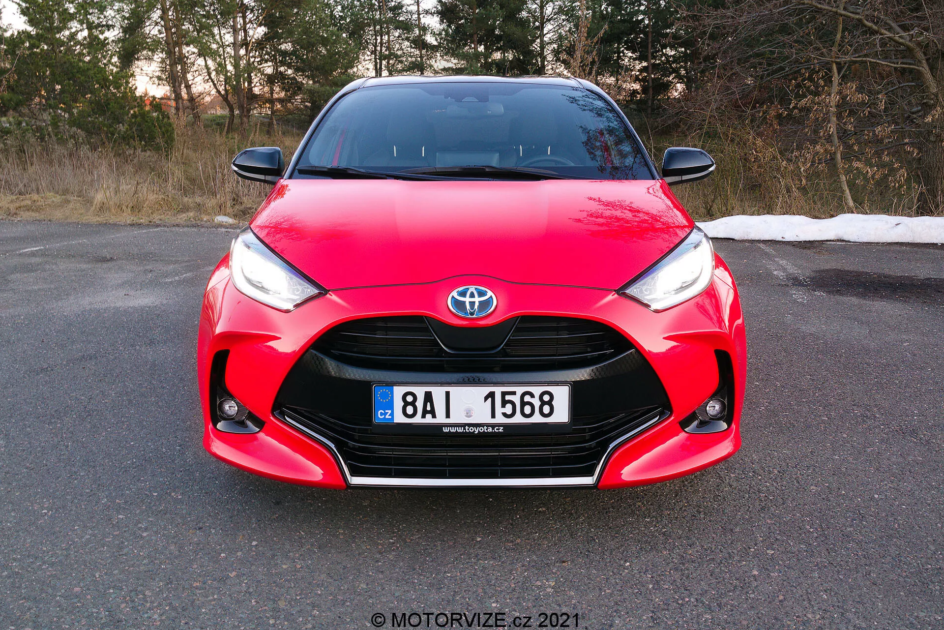 La imagen muestra una vista frontal de un Toyota Yaris 2020 rojo, tomada con un ángulo leve y enfocándose en la rejilla frontal, los faros y el área del capó, con la matrícula completamente visible. El coche presenta elementos de diseño distintivos, incluyendo el emblema de Toyota centrado en la rejilla, faros angulares agudos que se estrechan hacia la rejilla y curvas complejas en el capó, mostrando su diseño aerodinámico y deportivo. La foto, capturada durante el día antes del anochecer, sugiere que el coche está estacionado en una superficie plana en un entorno que podría ser finales de otoño o principios de primavera.