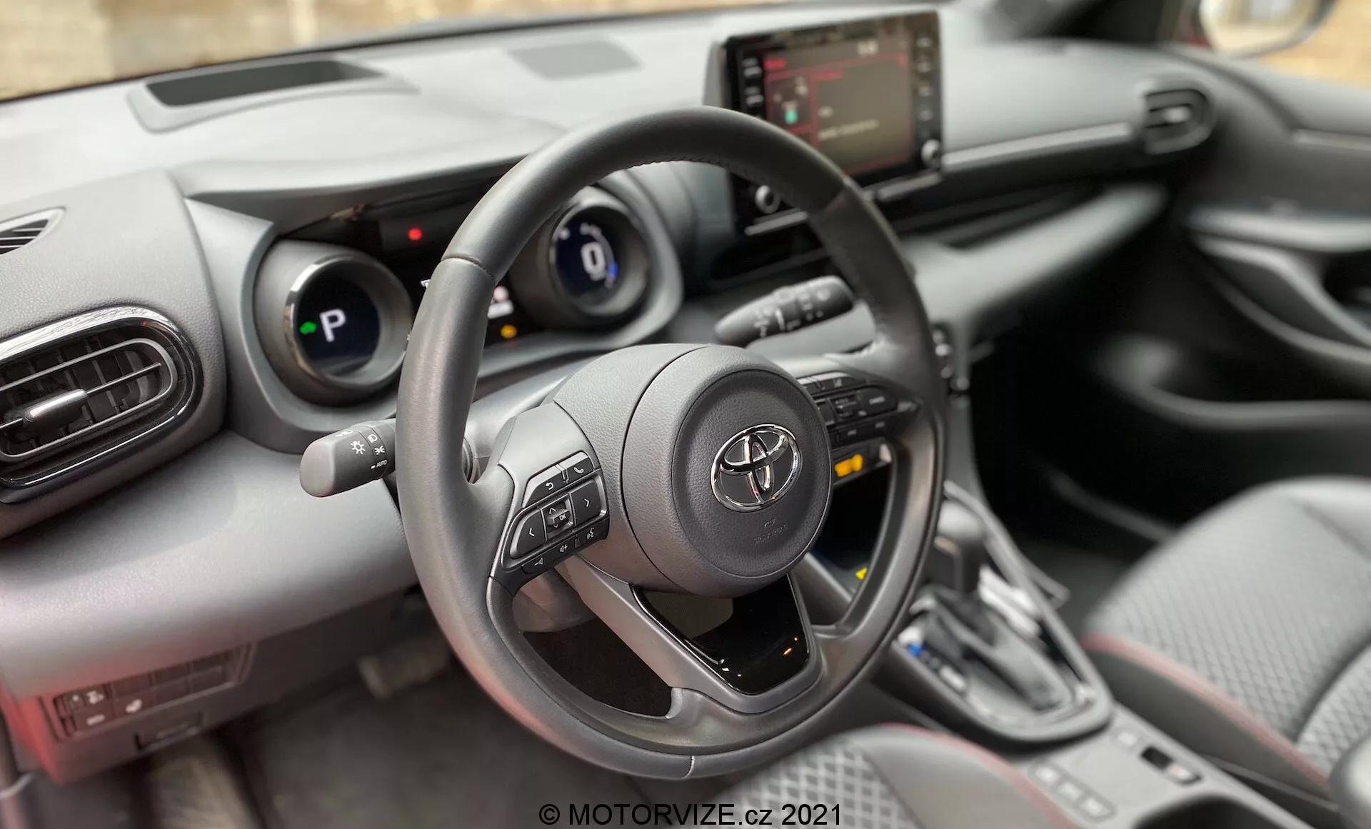 Esta imagen muestra el interior de un automóvil, enfocándose en la perspectiva del conductor. La toma es un primer plano, vista frontal del interior, con el ángulo de la cámara posicionado para capturar predominantemente el volante, el tablero y la consola central. La fotografía probablemente está tomada desde el asiento del conductor o posiblemente justo encima, ofreciendo un punto de vista similar al de una persona sentada detrás del volante. El volante es prominente en primer plano, centrado en el marco, y ocupa una parte sustancial de la imagen. Es de diseño de tres radios con controles multifunción montados en los radios izquierdo y derecho. Detrás del volante, el panel de instrumentos cuenta con una pantalla digital flanqueada por indicadores analógicos. A la derecha del volante, la consola central incluye una pantalla táctil de infoentretenimiento, panel de control climático y selector de marchas, indicando un vehículo moderno con funciones interactivas.