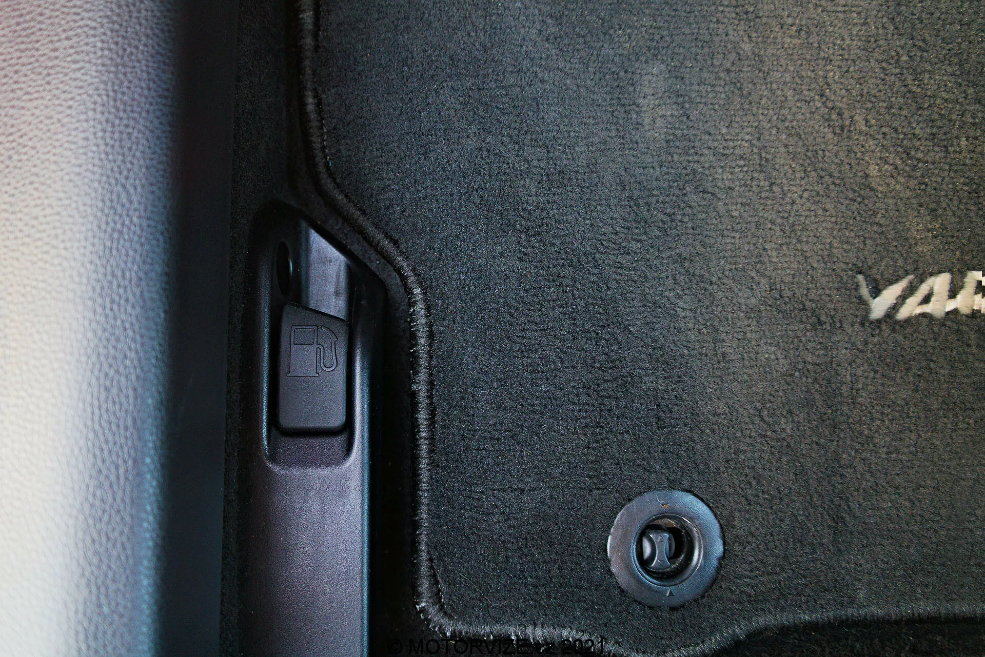 Vista de cerca del interior del Toyota Yaris 2020, capturando el lado izquierdo inferior cerca de la puerta. Se pueden ver una alfombra de color oscuro con texto bordado parcial, un gancho de retención circular metálico, el umbral de la puerta de plástico con una moldura protectora, una palanca de apertura del maletero que sugiere el lado del conductor, y un esquema de colores en dos tonos con texturas contrastantes de alfombra y plástico liso.