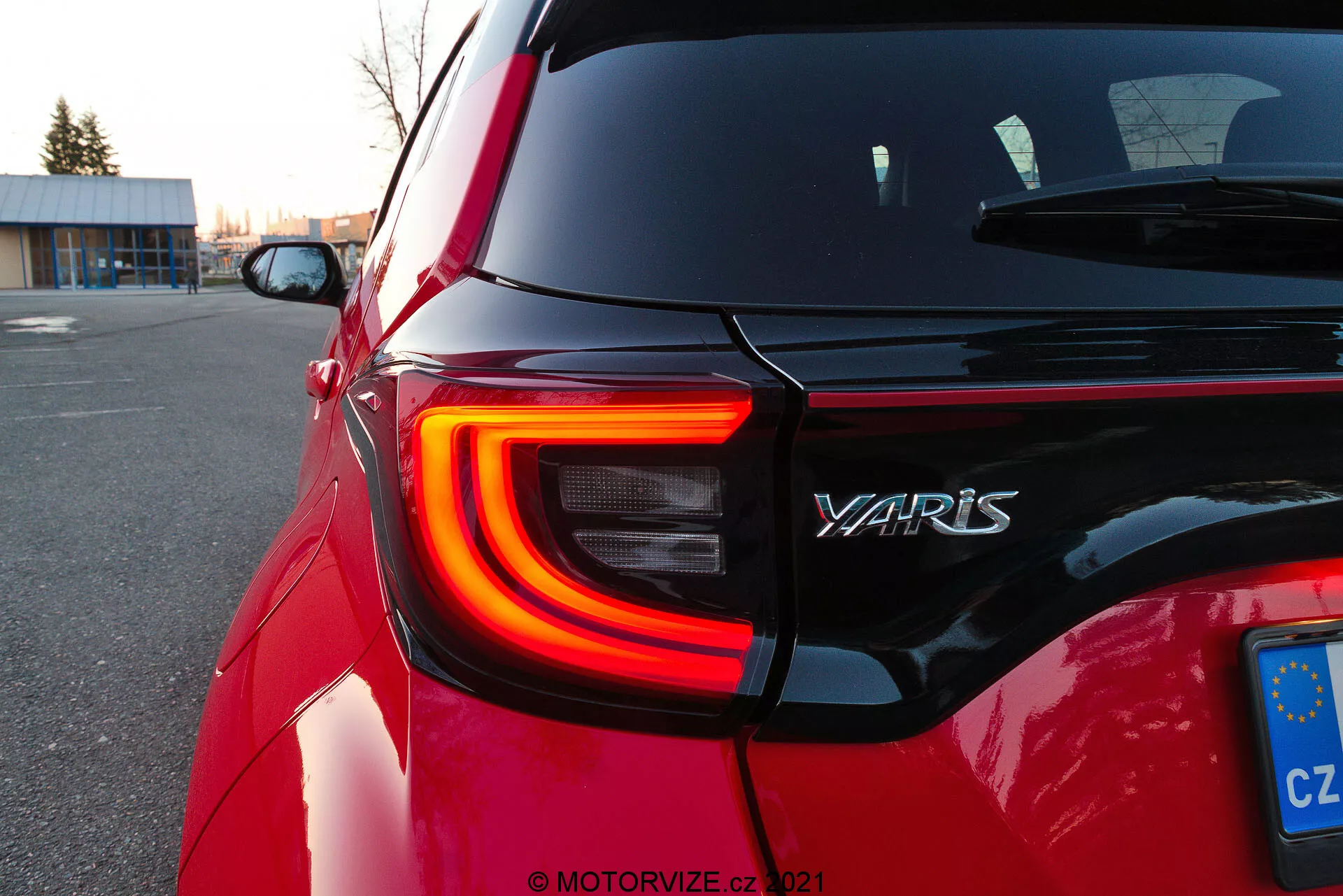 Imagen en primer plano de la sección trasera de un Toyota Yaris 2020 rojo, mostrando la luz trasera LED izquierda con su distintiva forma de C, el nombre del modelo 'YARIS' en el portón trasero y una placa de la Unión Europea de la República Checa. El ángulo oblicuo de la toma destaca los contornos y la reflectividad de la pintura del coche durante el crepúsculo en un aparcamiento, con fondos desenfocados que centran la atención en los detalles de diseño.