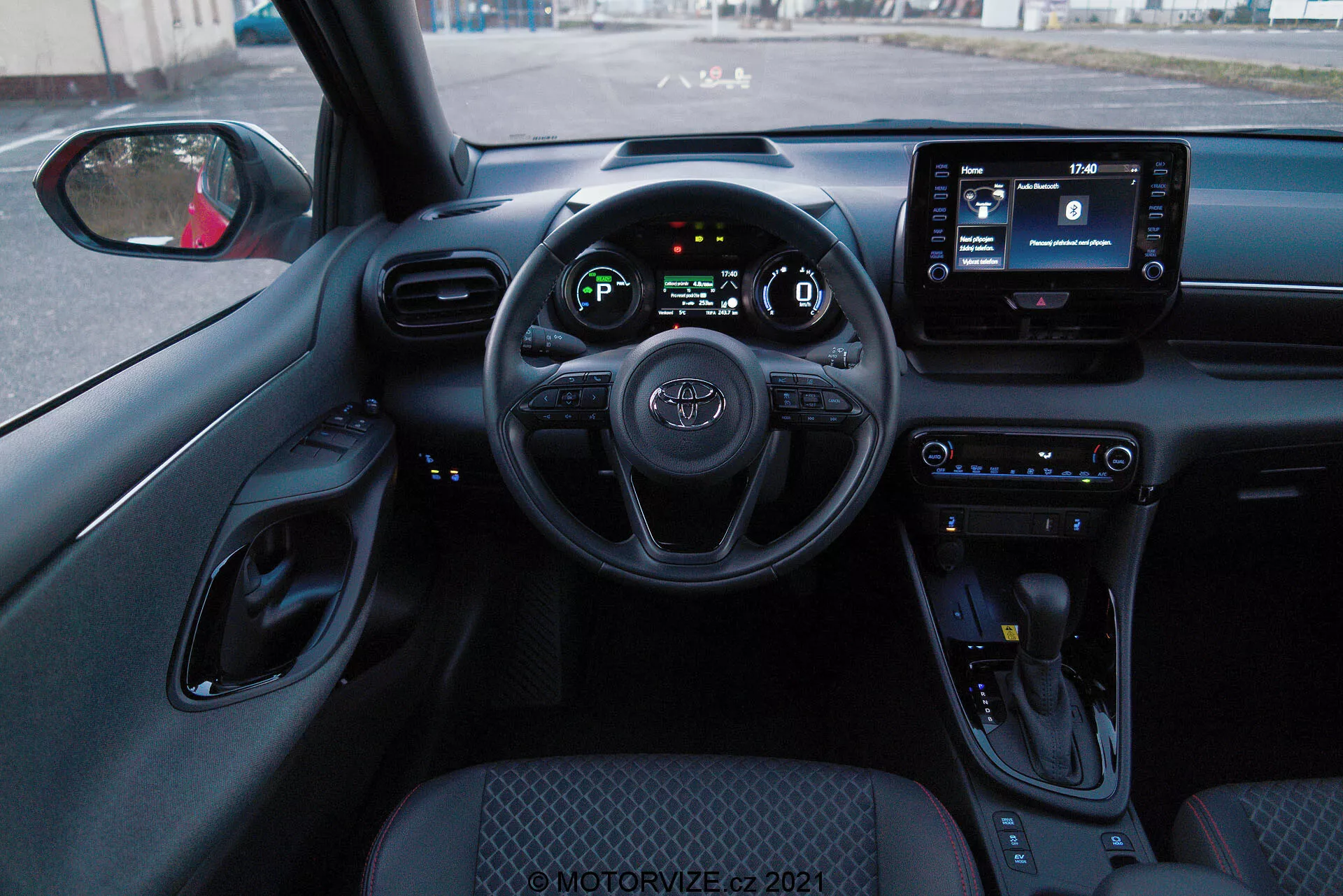 Perspectiva del interior desde el asiento del conductor de un Toyota Yaris 2020, mostrando el volante con controles montados, el panel de instrumentos digital y una pantalla de infoentretenimiento. El interior presenta botones de control de clima, una palanca de cambios y un esquema de color predominantemente negro con superficies texturizadas y acentos cromados.