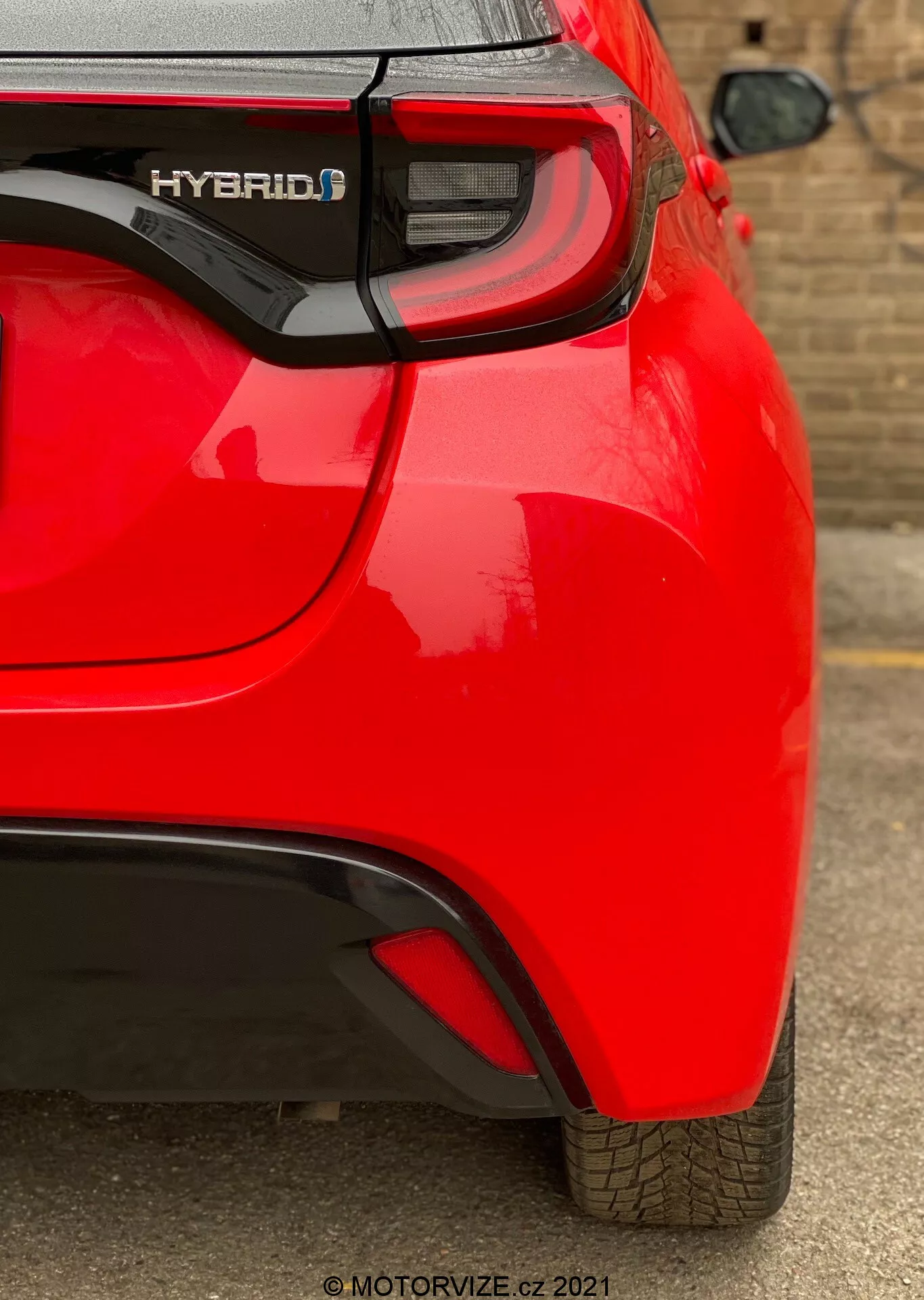 Vista trasera en ángulo de un coche híbrido Toyota Yaris 2020 rojo, enfocándose en el cuarto trasero derecho. Captura características como la insignia 'HYBRID', el clúster de luces traseras LED modernas, el sutil pliegue aerodinámico en el parachoques trasero y un sensor de estacionamiento visible. La pintura brillante y el dibujo de la banda de rodadura bien mantenido indican un vehículo nuevo o bien cuidado.