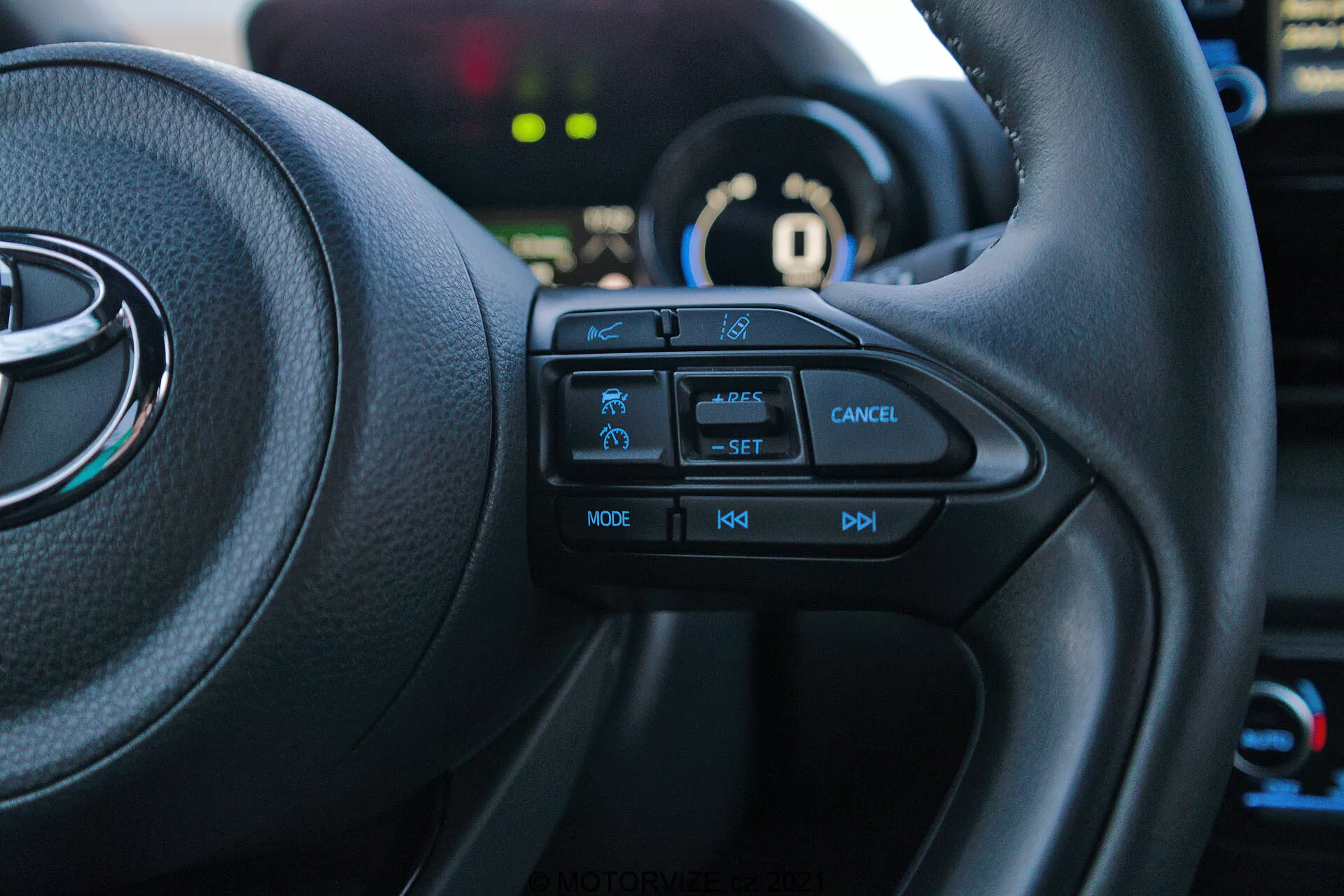 Imagen del interior de un Toyota Yaris 2020 desde el asiento del conductor, enfocada en el lado izquierdo del volante multifuncional. Los botones táctiles, incluidos el icono de la función de seguridad, control de crucero con etiqueta 'SET', botón de cancelación, 'MODE' para multimedia y controles de volumen '+', '-', están claramente visibles. El vehículo está en modo 'P' de aparcamiento, con el cuadro de instrumentos y elementos de fondo ligeramente desenfocados.