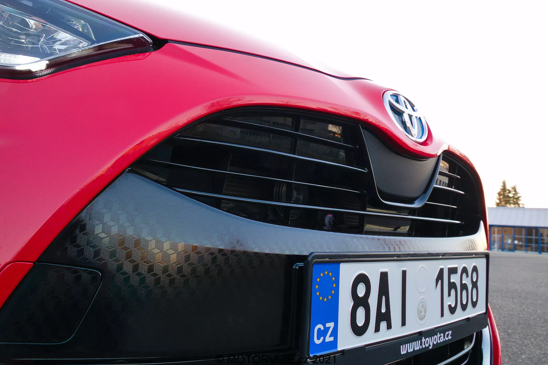 Imagen de primer plano de un Toyota Yaris 2020 rojo enfocándose en el lado delantero izquierdo, mostrando el capó, faro moderno con tecnología LED o HID, parrilla con emblema Toyota y patrón de panal, pintura roja brillante y parachoques delantero parcialmente visible con entrada de aire inferior. La matrícula de la Unión Europea indica registro en la República Checa.