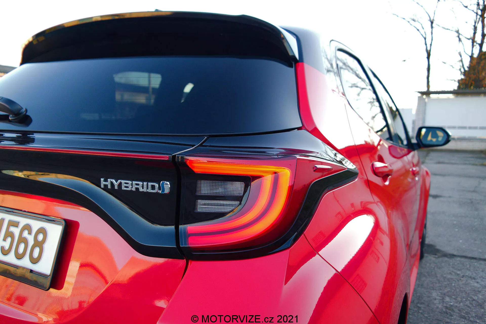 Imagen en primer plano y angulada de la sección trasera de un Toyota Yaris 2020 rojo, destacando el diseño moderno de la luz trasera LED con líneas curvas, la insignia HYBRID, el reflector integrado en el parachoques trasero, el limpiaparabrisas trasero, el alerón del techo, las líneas de calefacción del desempañador de la luneta trasera, la pintura roja reflectante y la zona de la matrícula parcialmente desenfocada con una cámara de visión trasera.