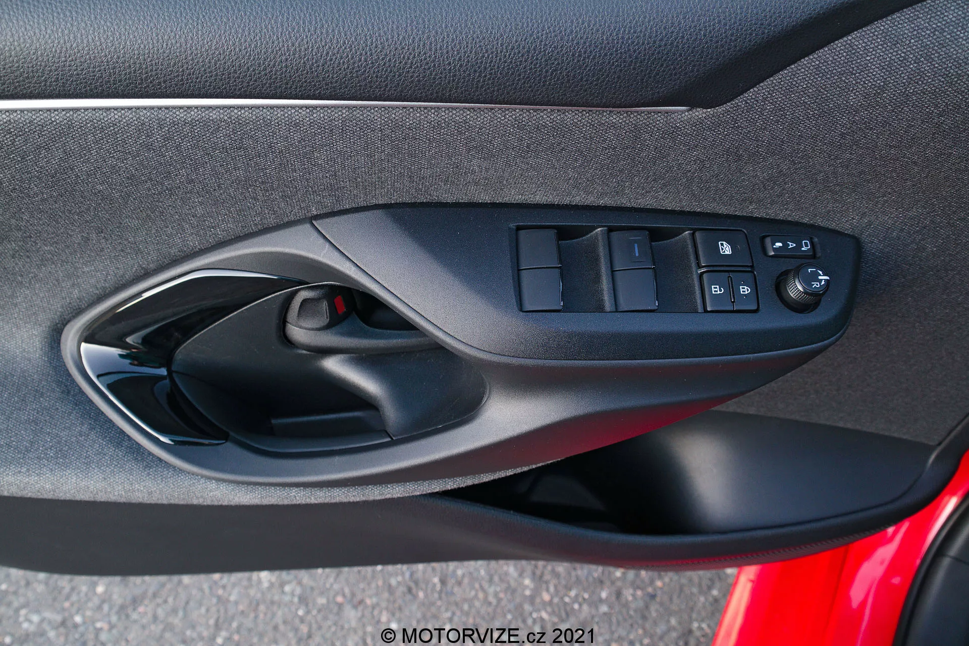 Panel interior de la puerta de un Toyota Yaris 2020 que se centra en los controles de la puerta del lado del conductor, diseñados ergonómicamente, incluidos los interruptores de las ventanas eléctricas, el botón de bloqueo de la puerta y los controles ajustables eléctricamente de los espejos, capturado desde la perspectiva de un ocupante dentro del vehículo. La foto resalta la mezcla de materiales suaves de tela o compuestos, el esquema de color negro y una rejilla de altavoz visible como parte del sistema de audio del coche.
