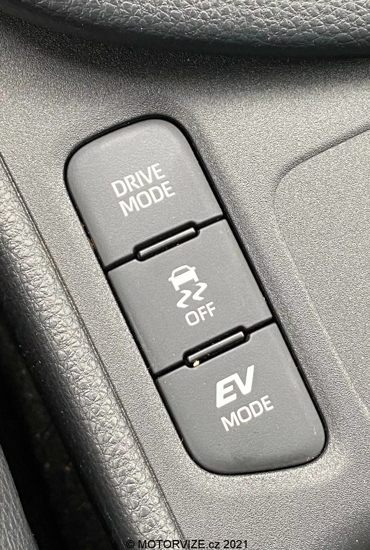 Toma de cerca del interior de un Toyota Yaris 2020, enfocando en tres botones de control alineados verticalmente relacionados con los modos de conducción. De arriba abajo: 'DRIVE MODE' con un icono de carretera sinuosa, 'Traction Control OFF' con un icono de coche derrapando y 'EV MODE', indicando la función de vehículo eléctrico. Los botones están montados en una superficie texturizada, probablemente parte de un panel de puerta o consola central. La imagen está enfocada nítidamente en los botones, con las texturas circundantes ligeramente desenfocadas, iluminada uniformemente por luz diurna para una visibilidad clara.
