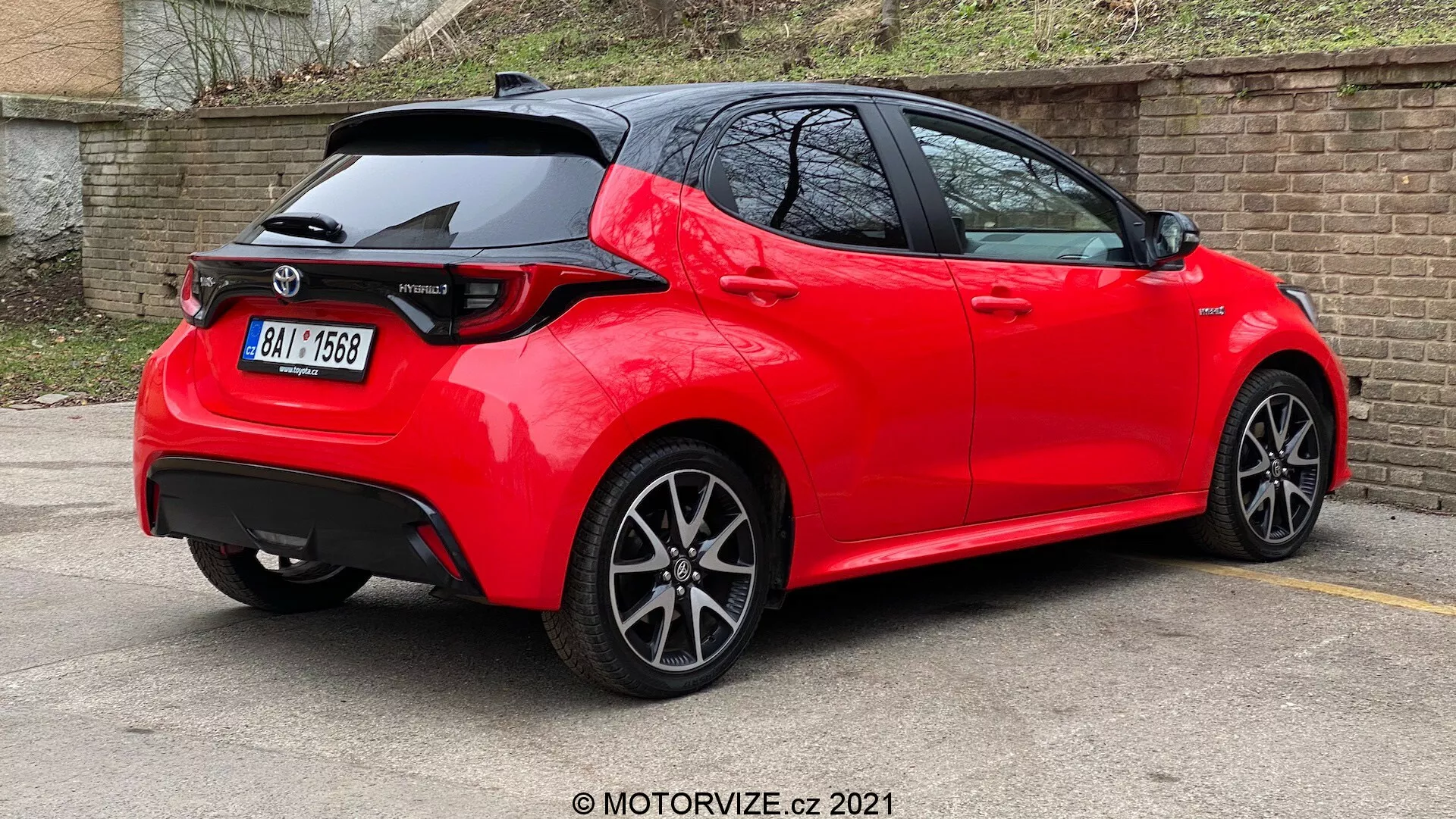Vista trasera de tres cuartos de un automóvil hatchback moderno de color rojo brillante, con líneas y pliegues definidos, un alerón en el techo, luces traseras LED, antena de aleta de tiburón, ruedas con múltiples radios y ubicado en un estacionamiento con pintura reflectante y bien cuidada, sugiriendo un modelo reciente o bien mantenido en una versión deportiva.
