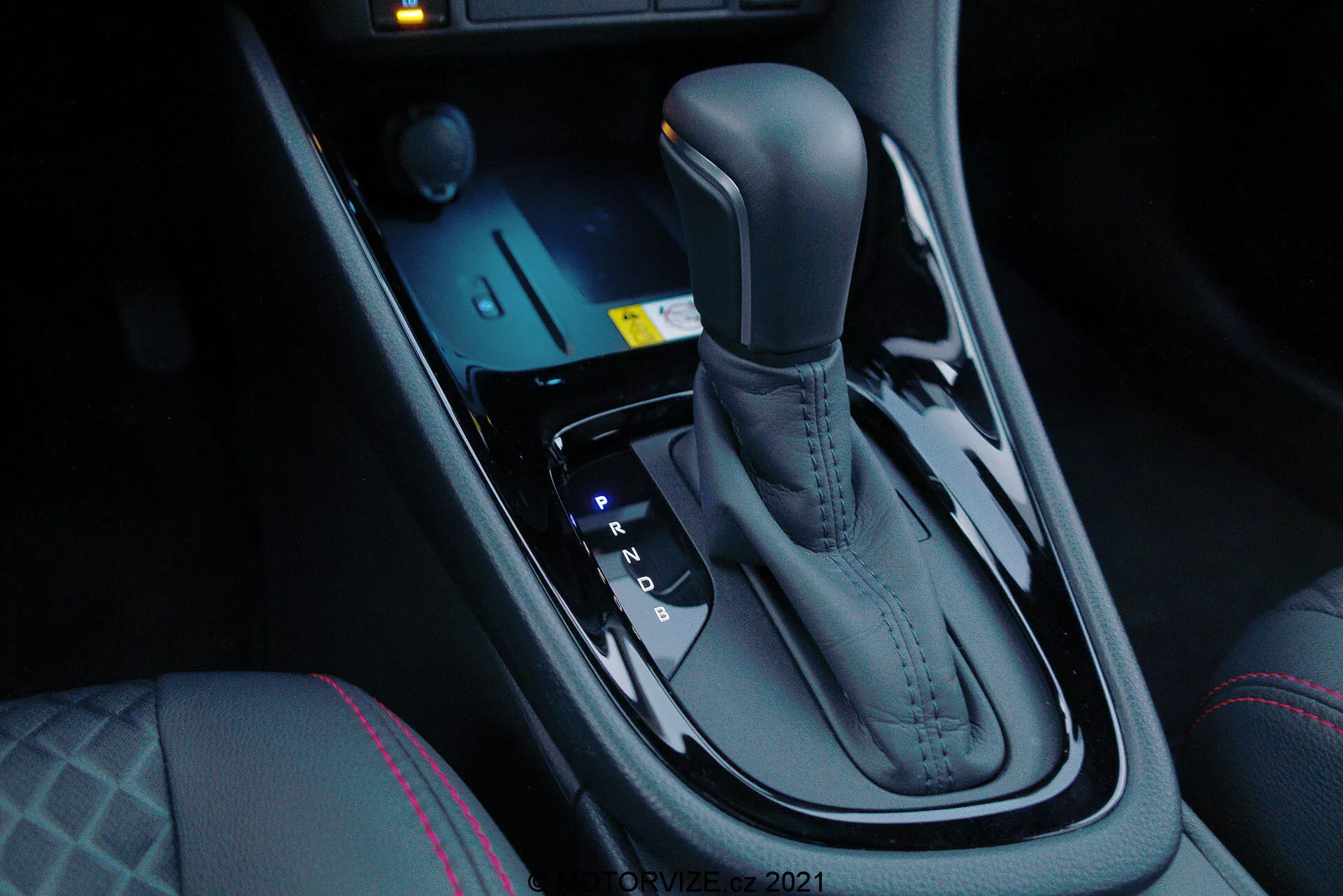 Vista de primer plano del selector de cambios automático del Toyota Yaris 2020, visto desde el lado del pasajero delantero en un ángulo descendente, destacando el acabado metálico elegante y el diseño ergonómico. El selector de cambios está situado en el centro entre los asientos del conductor y del pasajero delantero e incluye posiciones Park, Reverse, Neutral y Drive, así como opciones de cambio manual (+ y -). Delante del selector se encuentran controles electrónicos para modos de conducción y control de tracción, junto con un sistema de arranque por pulsador iluminado en azul. La consola central oscura y brillante añade un toque de lujo moderno, complementado por los controles de calefacción de los asientos y las costuras distintivas en el asiento del conductor, lo que indica mayor confort y dinámicas de conducción avanzadas.