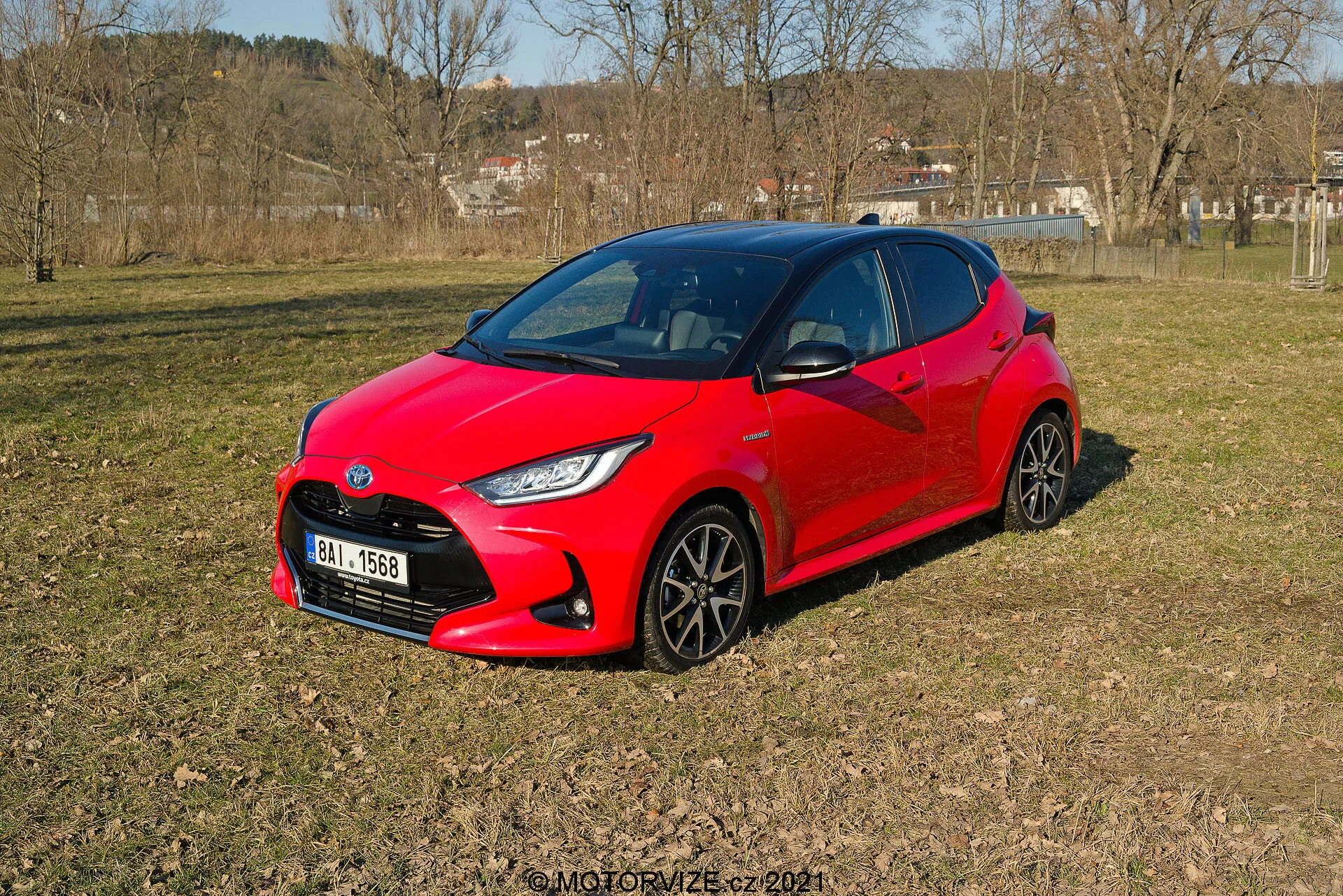 La imagen muestra un Toyota Yaris 2020 rojo vibrante, descansando en un terreno desigual y con hierba en un área semi-rural, visto desde una perspectiva de tres cuartos. El diseño moderno y aerodinámico del coche presenta ángulos agudos alrededor de los faros y una apariencia compacta y deportiva. Se pueden ver las ruedas oscuras de múltiples radios y los faros LED estilizados. Las ventanas laterales y el parabrisas delantero ofrecen un oscuro vistazo al interior, lo que sugiere posibles sistemas avanzados de automoción.