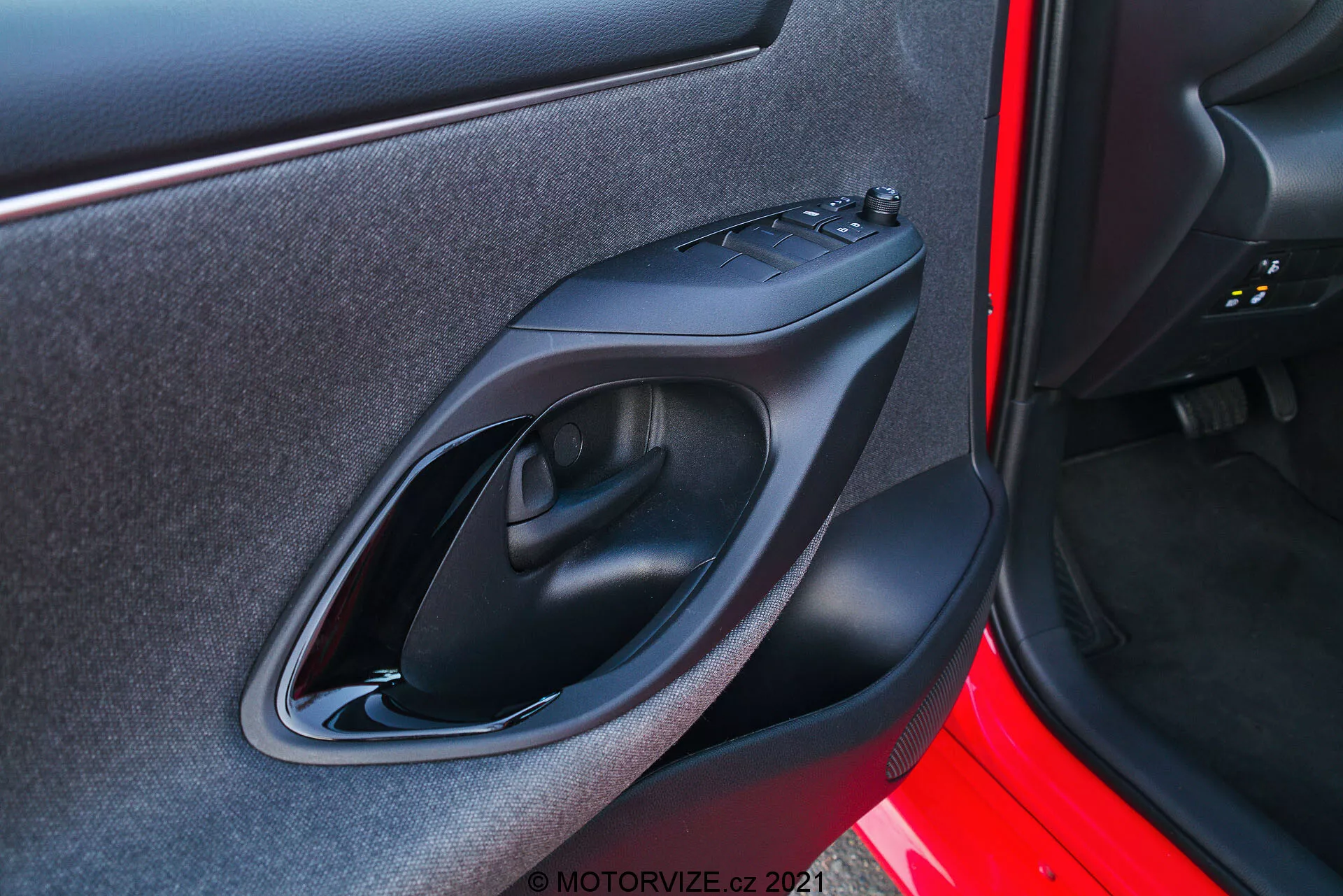 Primer plano del panel interior de la puerta de un Toyota Yaris 2020 tomado desde una perspectiva exterior. La imagen se centra en el panel de la puerta del lado derecho con características de diseño y tecnología como un tirador de puerta de plástico negro mate, panel de control de ventanas eléctricas, reposabrazos contorneado ergonómicamente y un pequeño bolsillo de almacenamiento. El exterior de la puerta está pintado en un color rojo vibrante.