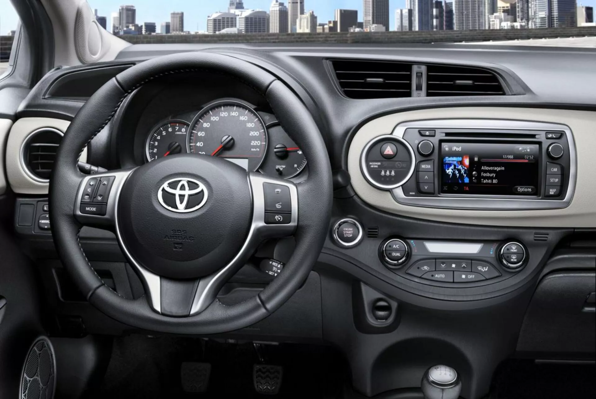 Vista interior del Toyota Yaris 2014 desde la perspectiva del conductor destacando el volante de tres radios con emblema de Toyota, controles montados para audio y teléfono manos libres, velocímetro y tacómetro analógicos con una pantalla digital, y un sistema de infoentretenimiento moderno con botones de control del clima en el tablero