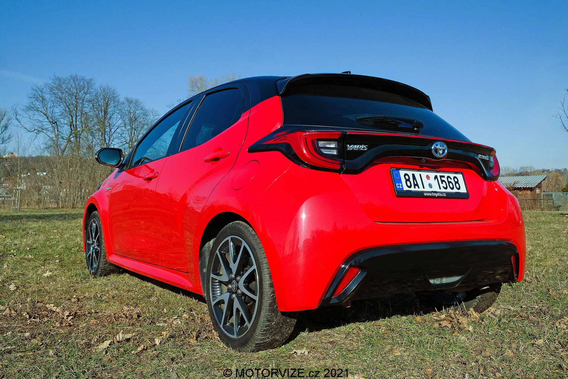 Vista trasera de tres cuartos de un hatchback Toyota Yaris 2020 rojo brillante, visto desde un ángulo bajo, destacando las líneas de diseño y características clave como luces traseras LED, alerón trasero, antena tipo aleta de tiburón y llantas de aleación de múltiples radios con acabado oscuro. La foto captura el detalle de la parte trasera y parte del perfil lateral a la luz del día, mostrando su estética deportiva y elementos modernos.