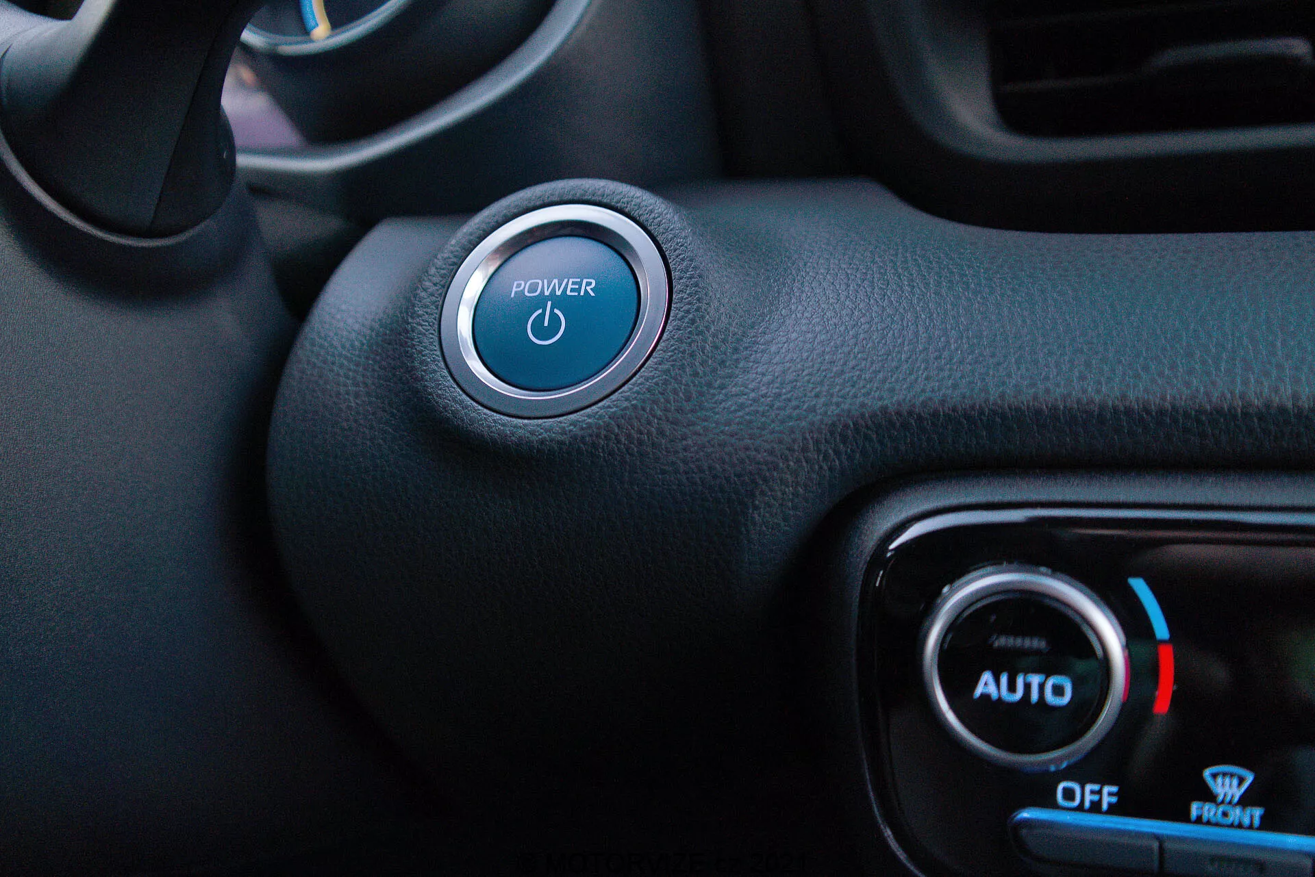 Vista interior del lado del conductor del Toyota Yaris 2020 destacando el botón de Start/Stop y el control de clima. El botón de encendido con un símbolo iluminado en azul es prominente en la parte superior izquierda, con un acento plateado. Abajo y a la derecha, los diales de control de clima muestran una luz azul y un icono de copo de nieve para temperatura fría, con etiquetas 'AUTO', 'OFF' y 'FRONT'. Una mezcla de materiales sintéticos texturizados y lisos en negro denota un estilo minimalista con características de arranque sin llave y control automático de clima.