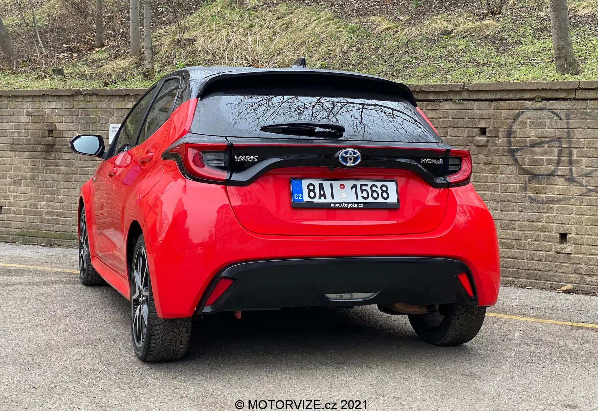 Una imagen de alta resolución de un Toyota Yaris 2020 hatchback rojo visto en un ángulo trasero de tres cuartos. El vehículo presenta luces traseras distintivas, un emblema central de Toyota sobre una probable cámara de visión trasera y la insignia del modelo 'Yaris' en la parte inferior izquierda de la puerta del maletero. Debajo del área de la matrícula hay una pieza de adorno negra que podría ser de plástico o fibra de carbono, con un pequeño alerón trasero que se extiende sobre la ventana trasera. El coche, aparcado cerca de un bordillo en un camino pavimentado junto a una pared de ladrillo, sugiere un entorno urbano o residencial.