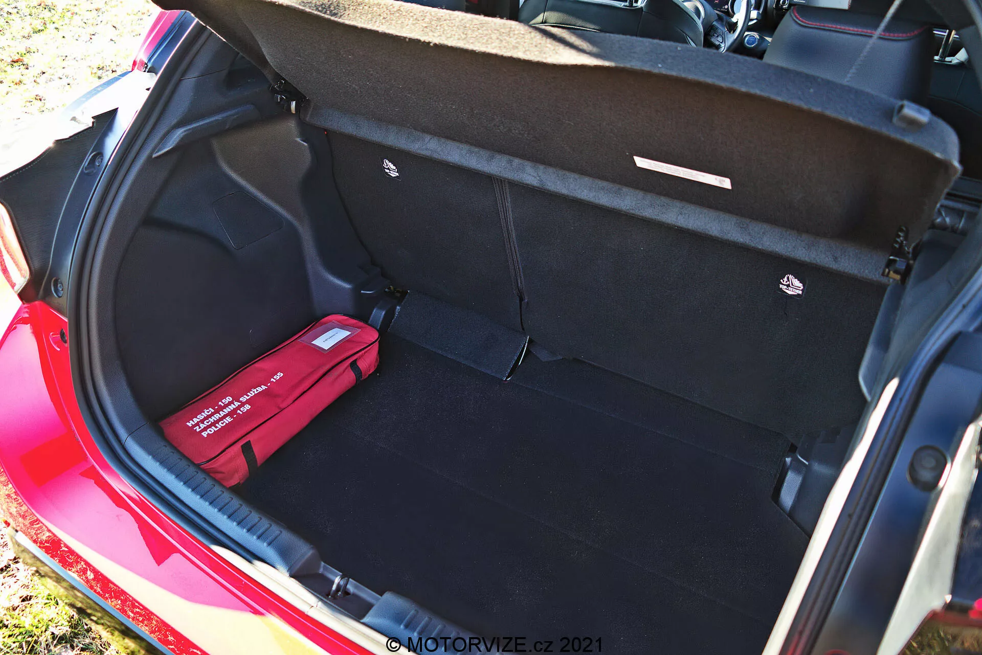 Maletero de un Toyota Yaris 2020 rojo visto desde un ángulo elevado, que muestra su interior limpio y espacioso con alfombrado negro, tapa del maletero abierta con manijas de tiro, un piso de carga plano, un gancho de carga a la derecha y un kit de primeros auxilios o de emergencia rojo a la izquierda. Esta fotografía diurna resalta la amplia capacidad de almacenamiento y características organizativas del vehículo.