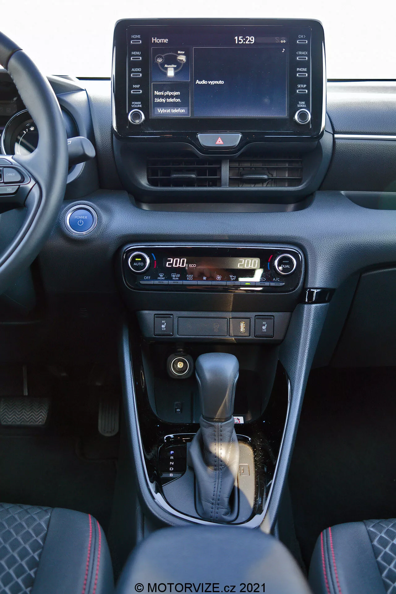 Vista interior de un Toyota Yaris 2020 enfocándose en la consola central y los elementos del tablero. La foto, tomada desde el asiento del pasajero delantero, muestra un sistema de infoentretenimiento digital con pantalla táctil que muestra una pantalla de inicio con la hora 15:29 y varias opciones de menú, ventilaciones centrales y controles fáciles de usar para el aire acondicionado y calefacción con una pantalla digital de temperatura ajustada a 20,0 grados. La consola central cuenta con una palanca de cambios envuelta en cuero con un acento plateado y un freno de mano manual. El interior está diseñado con tonos oscuros y acabados metálicos, ofreciendo una estética moderna y sofisticada.