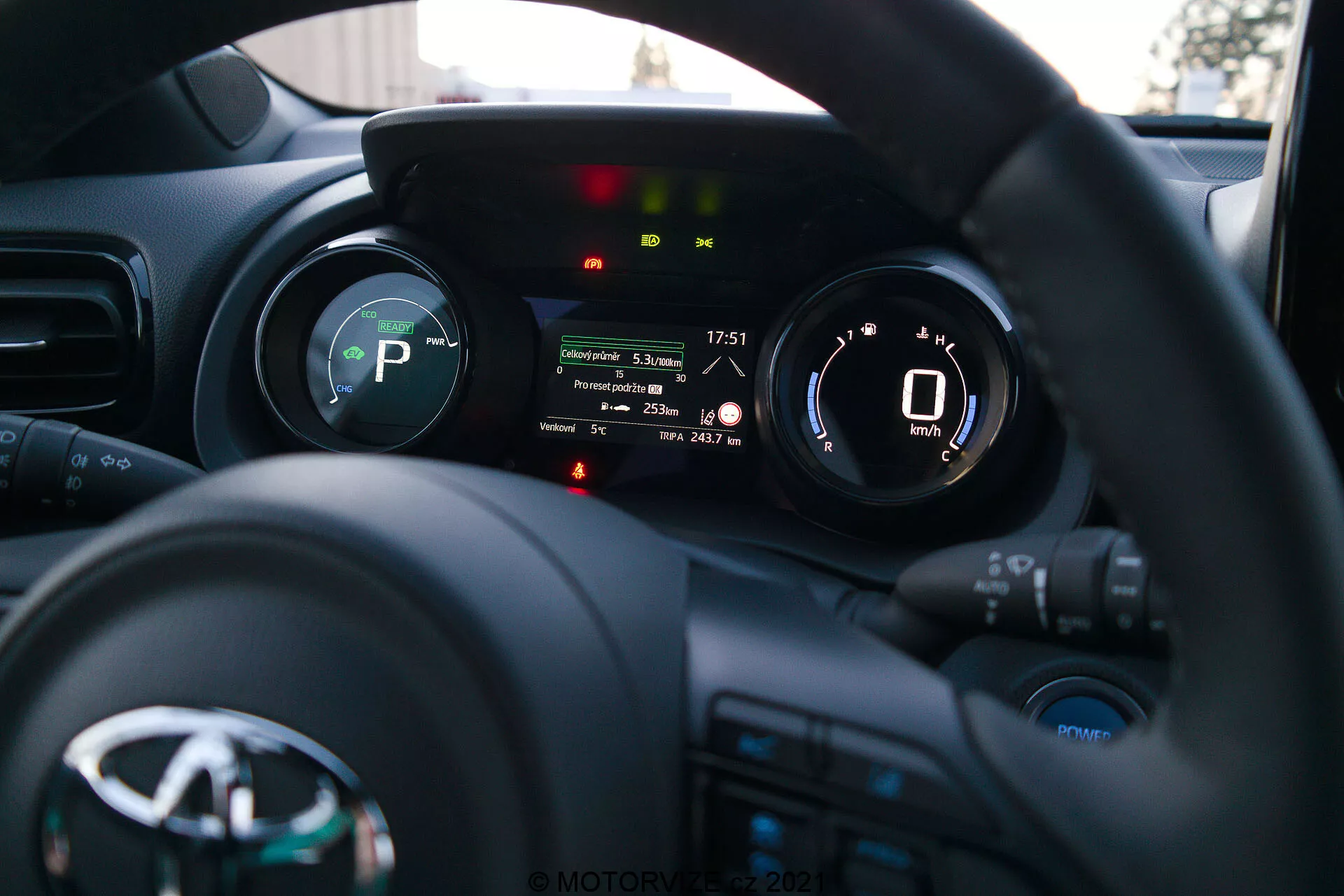 Una fotografía del interior del Toyota Yaris 2020 desde la perspectiva del conductor, enfocando el volante y el panel de instrumentos. El volante, con el logotipo del fabricante y varios botones de control para medios, comunicación y control de crucero, está en la parte inferior. El panel de instrumentos detrás del volante cuenta con una pantalla digital que muestra un velocímetro que marca 0 km/h e indicadores para la marcha (P de Park), recordatorio del cinturón de seguridad, alerta de puerta abierta y otras luces de estado del vehículo. La poca profundidad de campo resalta el panel de instrumentos, mientras que el tablero y el interior del coche están suavemente desenfocados en el fondo. La imagen, probablemente tomada a última hora de la tarde o a primera hora de la noche, muestra una instrumentación moderna y tecnología de interfaz del vehículo.