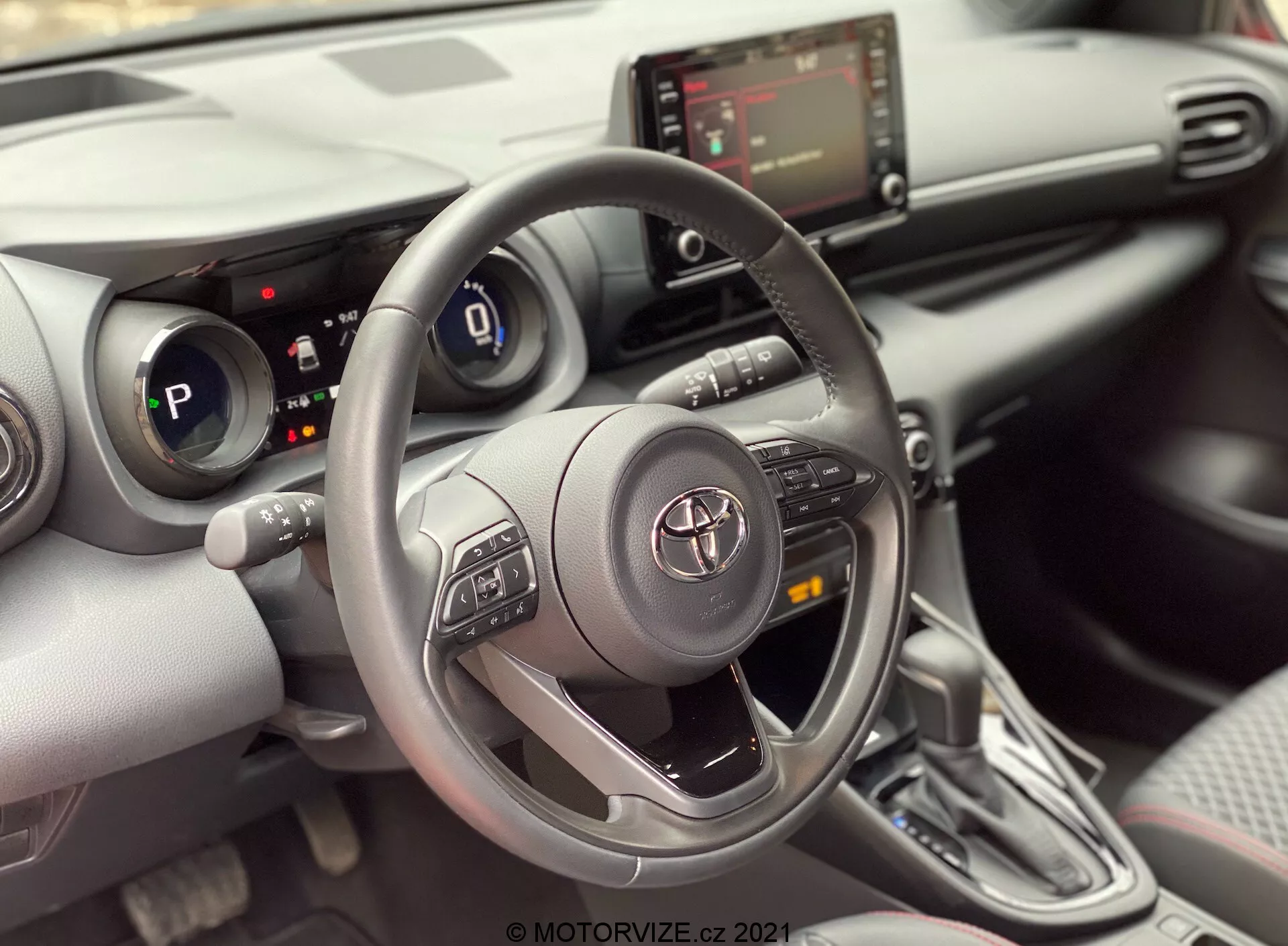 Vista interior de un Toyota Yaris 2020 desde el asiento del pasajero delantero, enfocándose en el área del conductor. La imagen destaca un volante de tres radios Toyota con botones de control para el audio, Bluetooth y control de crucero, con el emblema de Toyota en el centro. Detrás del volante, el cuadro de instrumentos digital es parcialmente visible indicando que el coche está en modo 'P' (aparcar). La consola central muestra la pantalla de infoentretenimiento y los controles del HVAC debajo de ella, junto con posibles puertos USB o auxiliares. La imagen también muestra una palanca de cambios automática con un diseño contemporáneo y una funda tipo cuero. El interior tiene un diseño moderno con una mezcla de materiales suaves al tacto y acentos plateados, junto con porta vasos o pequeños compartimientos de almacenamiento al lado de la palanca de cambios. Un panel de la puerta parcialmente visible revela un panel de control de ventanas, sugiriendo ventanas eléctricas.