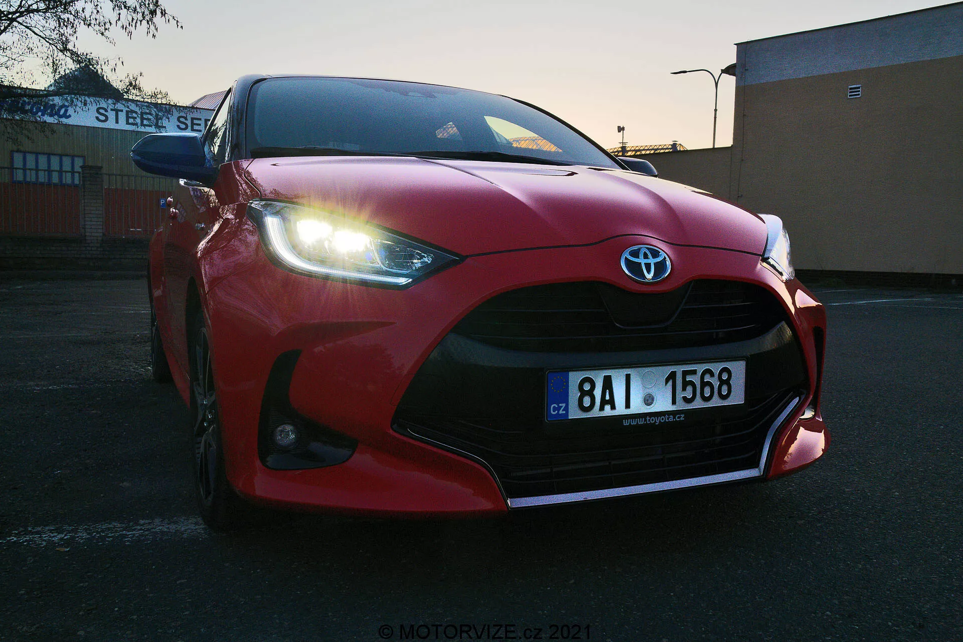 Vista de primer plano en ángulo bajo de un Toyota Yaris 2020 rojo estacionado al aire libre al amanecer o atardecer. Rejilla frontal, parachoques, capó aerodinámico esculpido, faros LED afilados y grandes entradas de aire destacando el emblema de Toyota. Matrícula europea visible, situado contra un fondo industrial o comercial con el edificio 'STEEL SERVICE'. Cielo despejado con luz en degradado resaltando el diseño deportivo del vehículo.