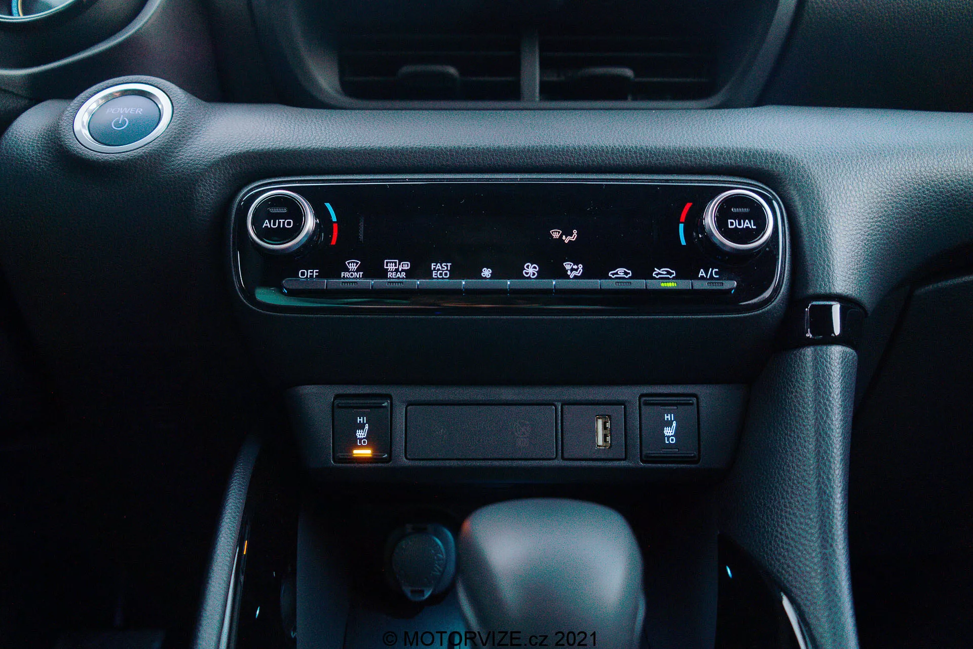 Vista de primer plano del tablero de instrumentos del Toyota Yaris 2020: La imagen destaca el sistema de control de clima con climatización automática de doble zona, con botones 'AUTO' y 'DUAL' para ajustes independientes de temperatura para el conductor y el pasajero. Debajo de los controles de clima, se ve un compartimiento de almacenamiento con un puerto USB y una posible toma de corriente de 12V. A continuación, se observan tres mandos: el central es la palanca de cambios en 'Parque', lo que indica una transmisión automática, un mando giratorio más pequeño a la izquierda probablemente para la selección del modo de conducción, y un botón en la derecha que podría ser un freno de mano electrónico. El material del tablero tiene un esquema de colores oscuros con un acabado texturizado premium, creando un entorno de cabina moderno y fácil de usar.