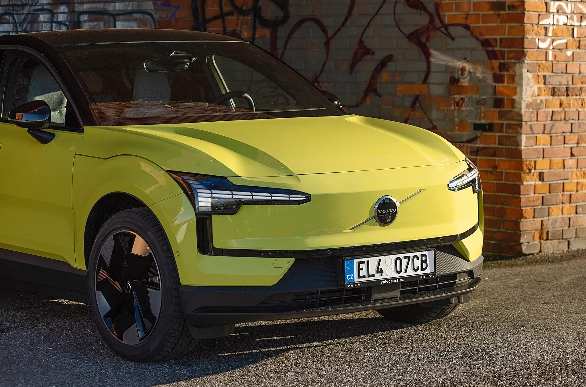 Coche eléctrico Volvo EX30 verde lima del 2024 aparcado frente a una pared de ladrillo con graffiti, mostrando diseño aerodinámico, faros LED modernos y ausencia de parrilla frontal