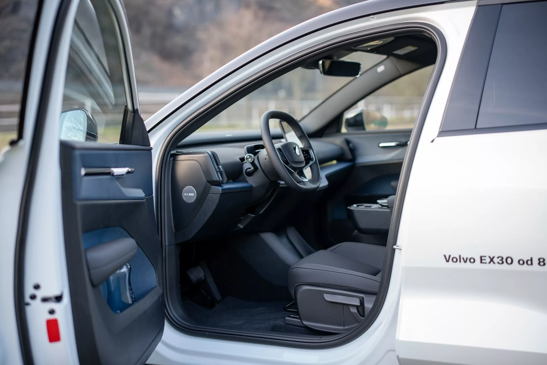 Vista interior de un Volvo EX30 2024 blanco, mostrando el asiento del conductor y el área del cockpit con la puerta delantera izquierda abierta que presenta controles de energía, y un vistazo del volante y parte del tablero