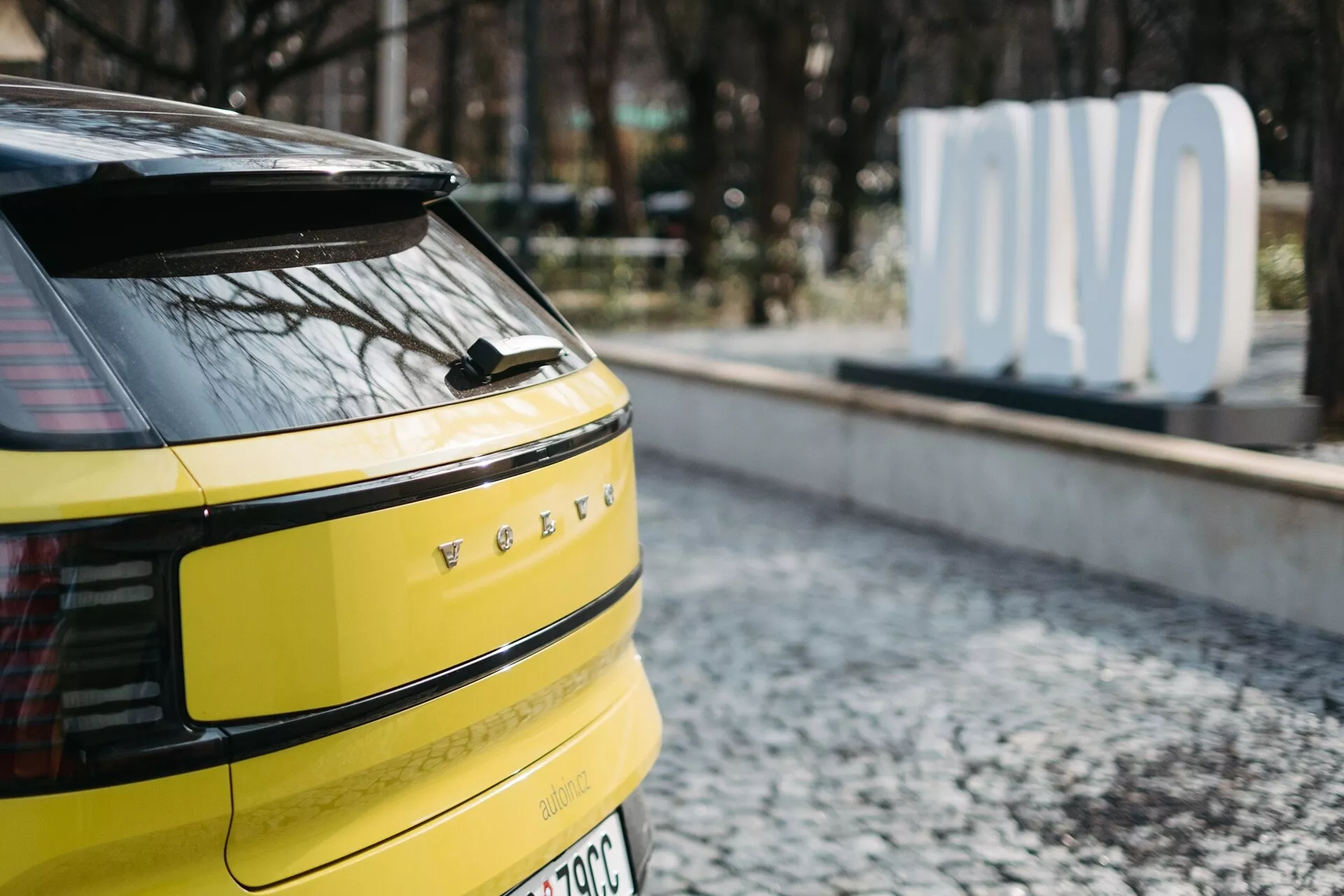 Primer plano de la sección trasera de un Volvo EX30 2024 amarillo brillante, con luces traseras LED horizontales, posible cámara o sensores traseros, limpiaparabrisas trasero único, distintivos de vehículo eléctrico o híbrido y antena tipo aleta de tiburón, estacionado sobre superficie de adoquines con un cartel de 'OLVO' desenfocado en el fondo