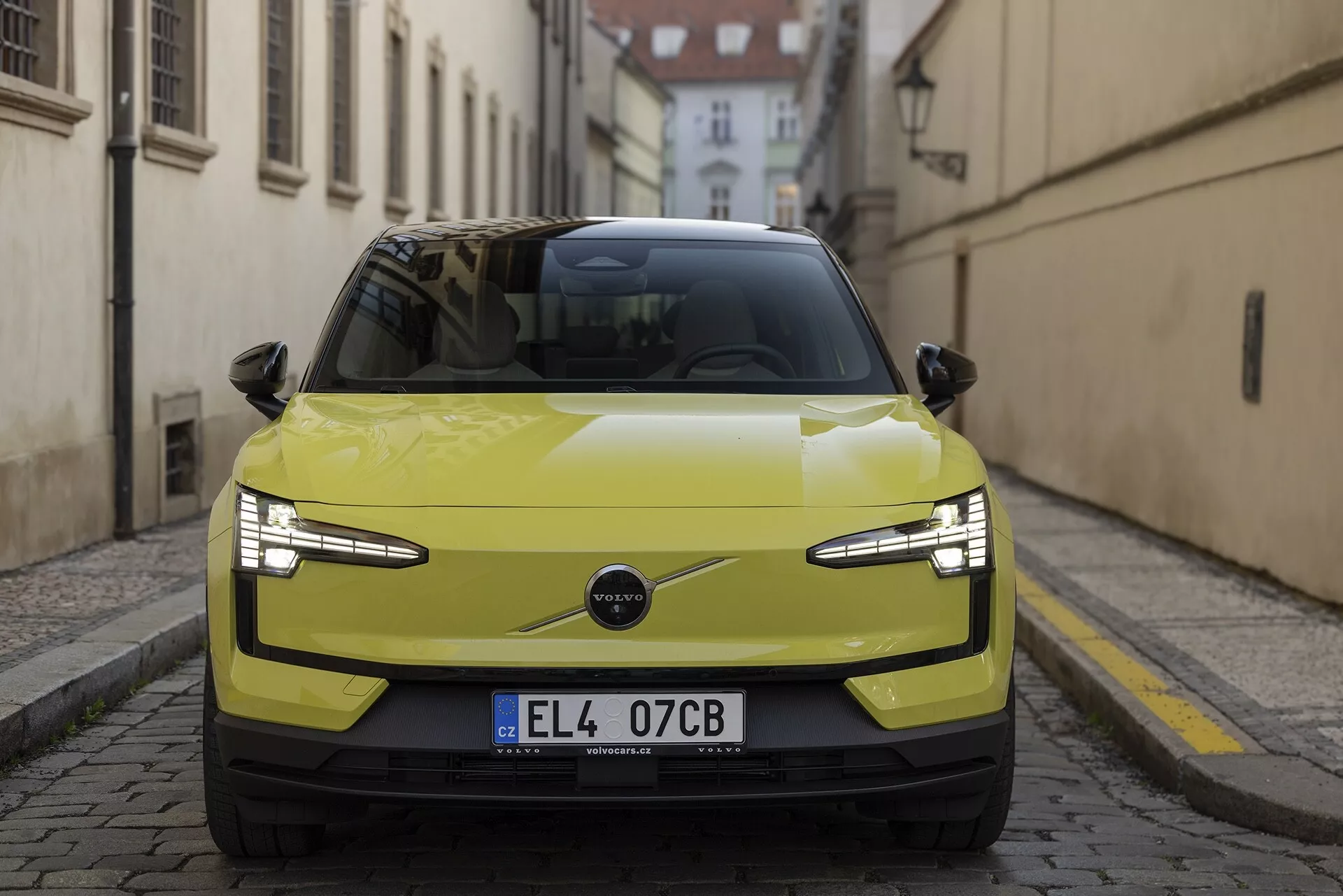 Vista frontal del Volvo EX30 2024 en color amarillo lima, destacando sus elegantes faros LED, rejilla minimalista indicativa de un tren motriz eléctrico y características tecnológicas avanzadas como sensores de estacionamiento y posible ADAS, contra una calle europea histórica