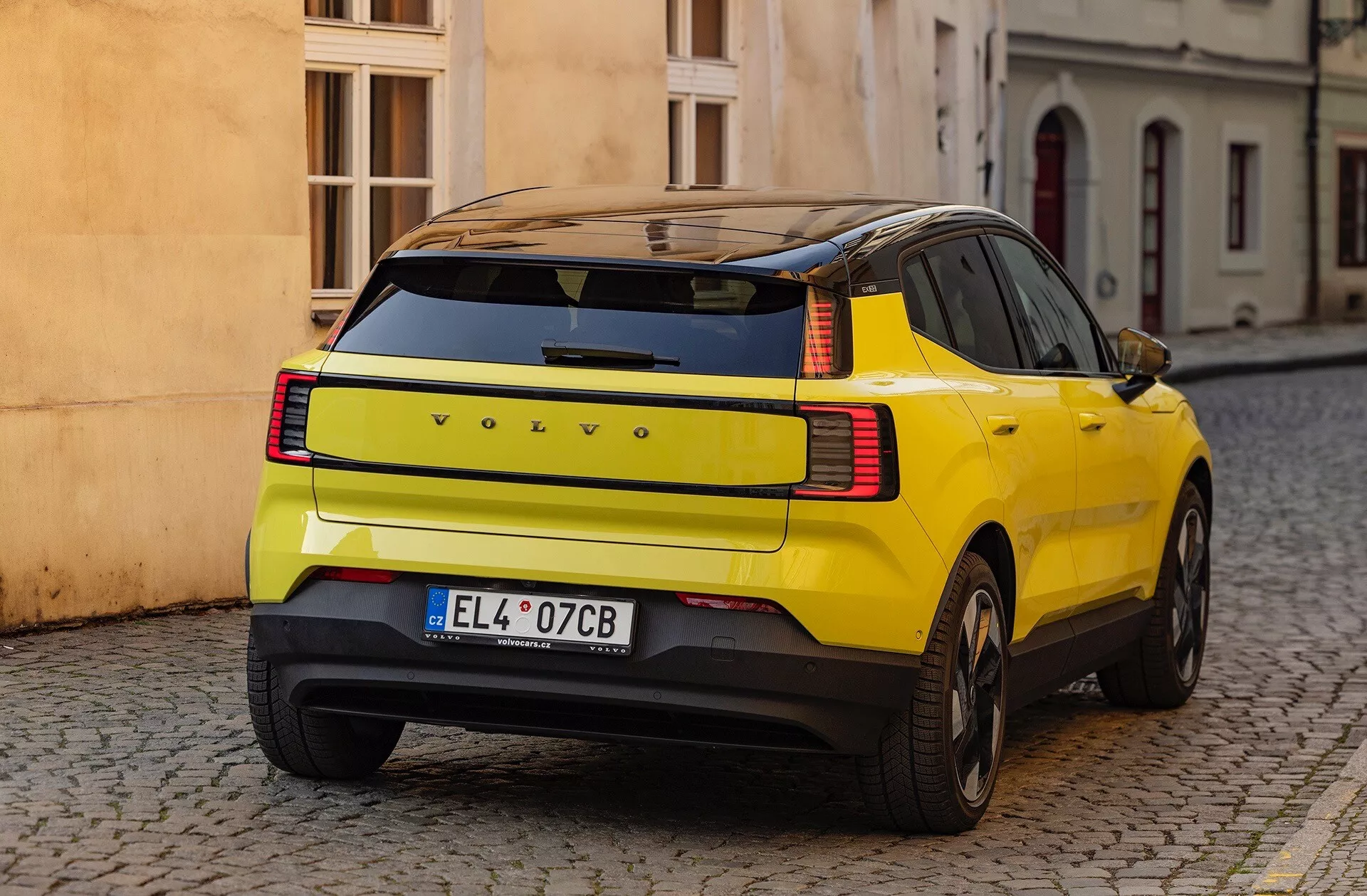 SUV amarillo Volvo EX30 2024 mostrando su distintiva curvatura del panel trasero, luces traseras LED y características de seguridad, aparcado en una calle adoquinada desde una vista trasera de tres cuartos