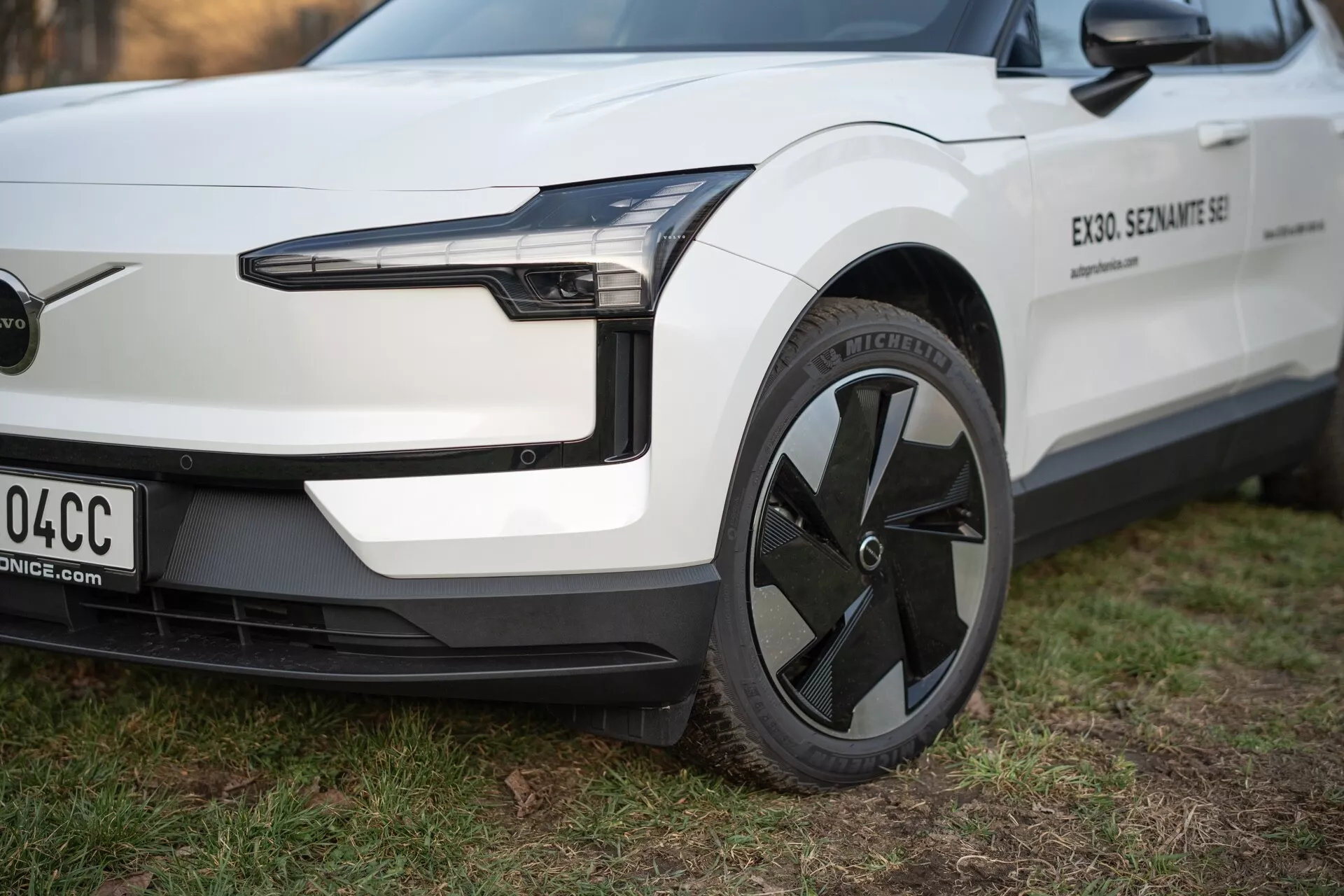Vista detallada del vehículo eléctrico Volvo EX30 2024 desde una perspectiva de tres cuartos, destacando su faro LED, diseño de rueda aerodinámico y parachoques delantero minimalista sin rejilla, en blanco con detalles en negro.