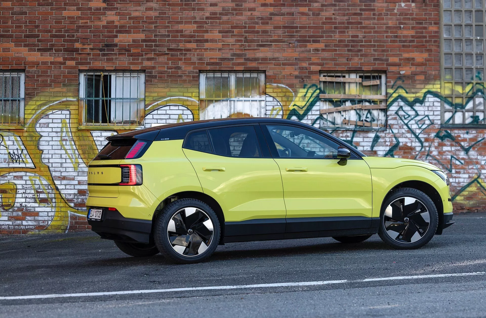 Imagen lateral de un SUV Volvo EX30 verde lima del 2024 estacionado junto al bordillo de una calle urbana con arte de grafiti de fondo, mostrando sus llantas deportivas negras de aleación, luces traseras LED y luz de freno alta montada.