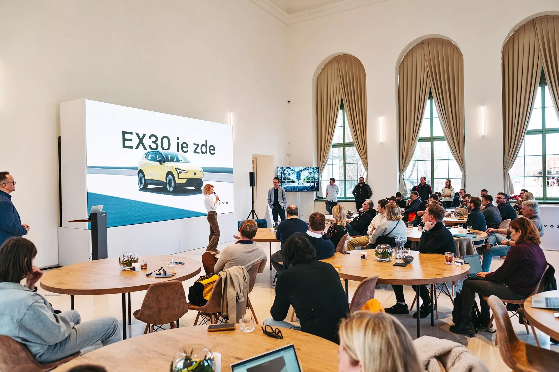 Presentación del vehículo eléctrico Volvo EX30 2024 en una sala espaciosa con techos altos y ventanas altas arqueadas, mostrando un EX30 amarillo desde una vista frontal de tres cuartos en una gran pantalla