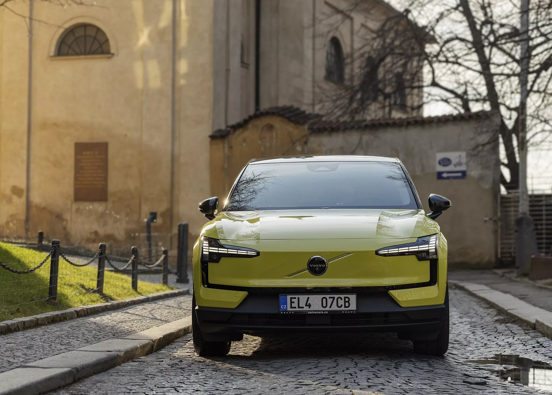 Coche eléctrico moderno Volvo EX30 2024 en color chartreuse aparcado en una calle de adoquines, visto desde una perspectiva frontal-derecha con faros LED y diseño aerodinámico del capó destacados contra un telón de fondo europeo histórico.