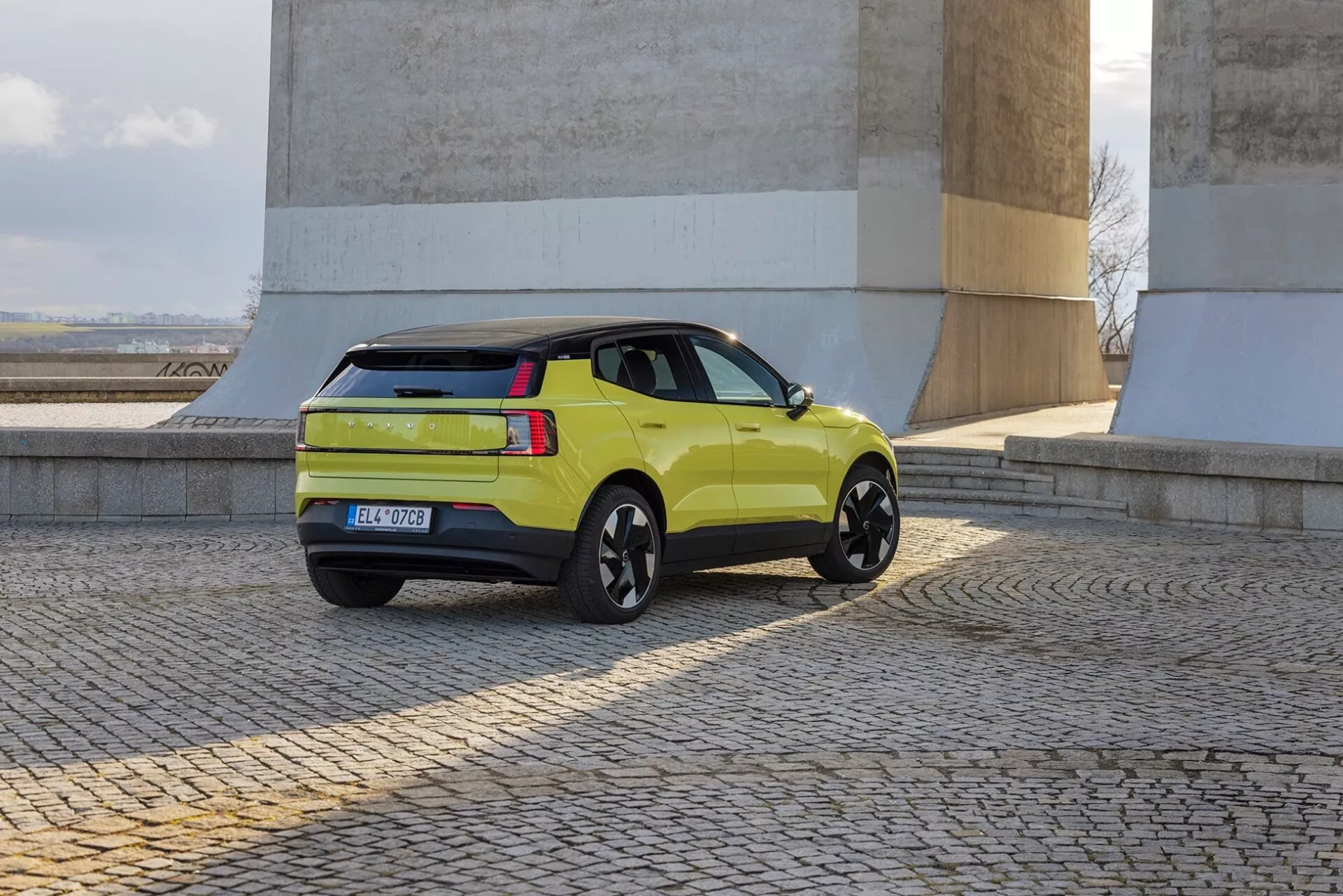 SUV verde lima Volvo EX30 2024 en vista trasera de tres cuartos mostrando perfil lateral, luces traseras, portón trasero y ruedas de aleación negras cerca de paredes de piedra sobre suelo adoquinado