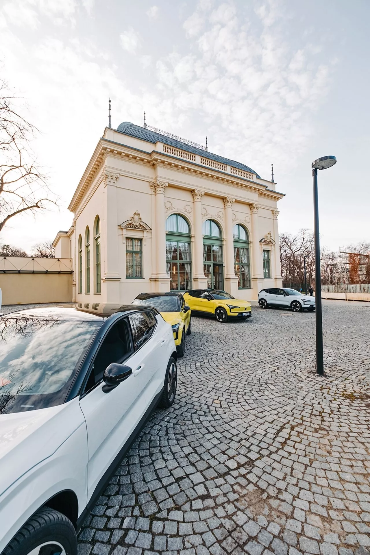 Escena callejera con tres coches y un edificio elegante al fondo, tomada desde una perspectiva de gran angular destacando un sedán contemporáneo blanco, un deportivo amarillo vibrante y un sedán o hatchback blanco a la luz del tardío atardecer