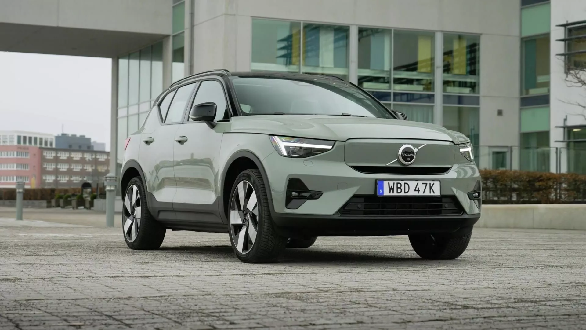 SUV eléctrico Volvo EX40 1ª generación del 2024 aparcado en pavimento con edificios urbanos al fondo, mostrando su fachada aerodinámica con parrilla cerrada, faros LED elegantes, grandes ruedas de aleación de múltiples radios, diseño moderno de carrocería con manijas de puertas al ras, señales de giro integradas en los espejos laterales, ventanas enmarcadas en cromo y tecnología avanzada de asistencia al conductor.
