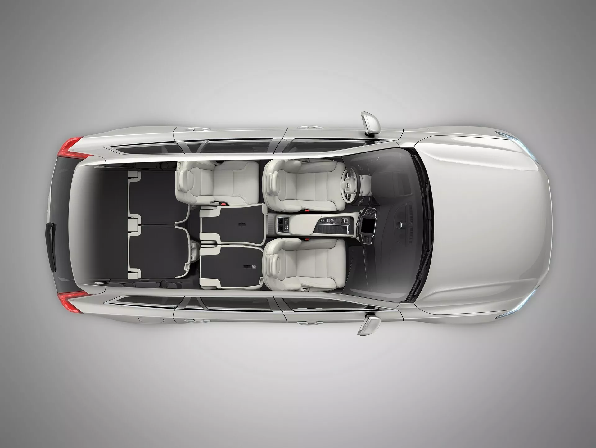 Vista de pájaro de un sedan Volvo XC90 de segunda generación del 2015 con techo transparente o digitalmente eliminado que muestra el interior. La imagen revela el capó largo del coche, cuatro puertas cerradas, cuatro ruedas y la configuración de asientos con dos asientos delanteros y un banco trasero para tres pasajeros. Consola central y palanca de cambios visibles. Se muestra el tablero de instrumentos con volante a la izquierda, pantalla y distribución de controles. Elementos de diseño como reposacabezas, espejos exteriores y el espacio y disposición interior son claramente visibles.