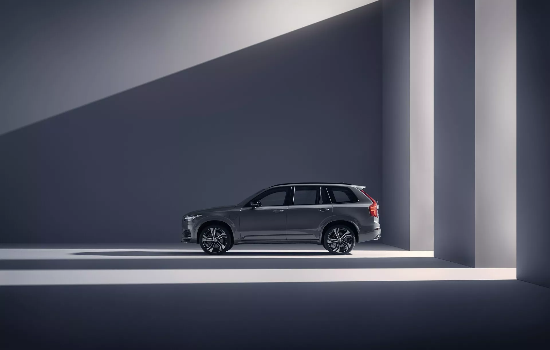 Perfil lateral de un Volvo XC90 2015 (2ª generación) en la categoría de SUV de lujo. La foto está tomada en un estudio o sala de exposición, mostrando las líneas de diseño del vehículo, manijas de puerta elegantes, ruedas de aleación de múltiples radios grandes y adornos cromados en las ventanas. Los rieles del techo sugieren utilidad junto con lujo. El vehículo está bien iluminado con una iluminación dramática que crea sombras geométricas, enfatizando su apariencia moderna y de alta gama. Las características avanzadas como la entrada sin llave se infieren a través de elementos de diseño.