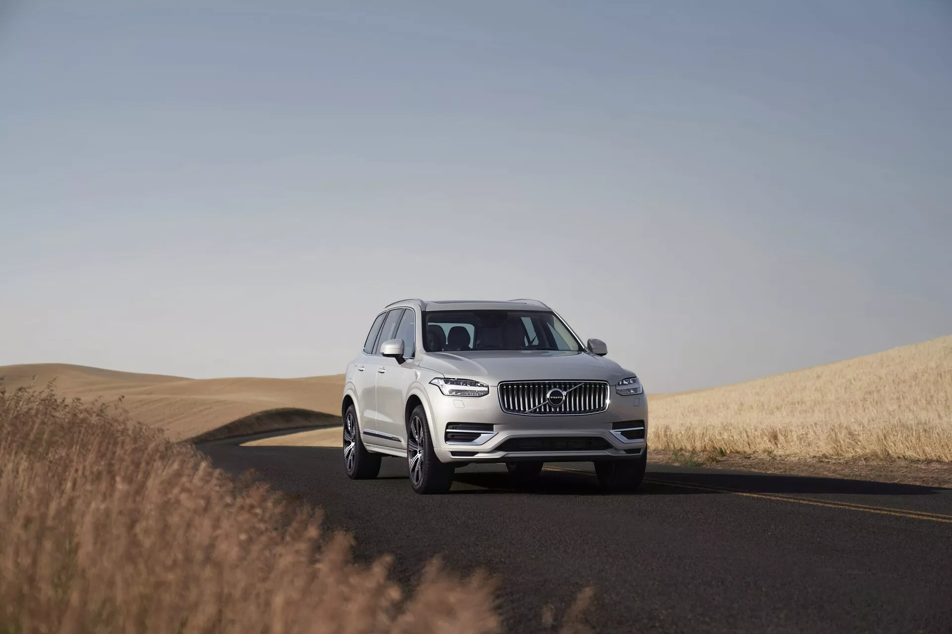 Un SUV Volvo XC90 2015 blanco en una carretera de dos carriles, capturado durante el día con el cielo despejado. La imagen muestra el vehículo desde un ángulo bajo a medio, destacando su imponente presencia y dimensiones. Posicionado en el centro del marco con una vista de tres cuartos, la foto captura detalles de diseño como la parrilla, los faros LED, el emblema y el parachoques delantero, indicando sistemas avanzados de asistencia al conductor. Las ruedas, las aleaciones y posiblemente el tamaño de los neumáticos son claramente visibles. El entorno sereno presenta una carretera que gira a la derecha y campos de trigo dorado, destacando la idoneidad del vehículo para viajes largos en paisajes diversos.
