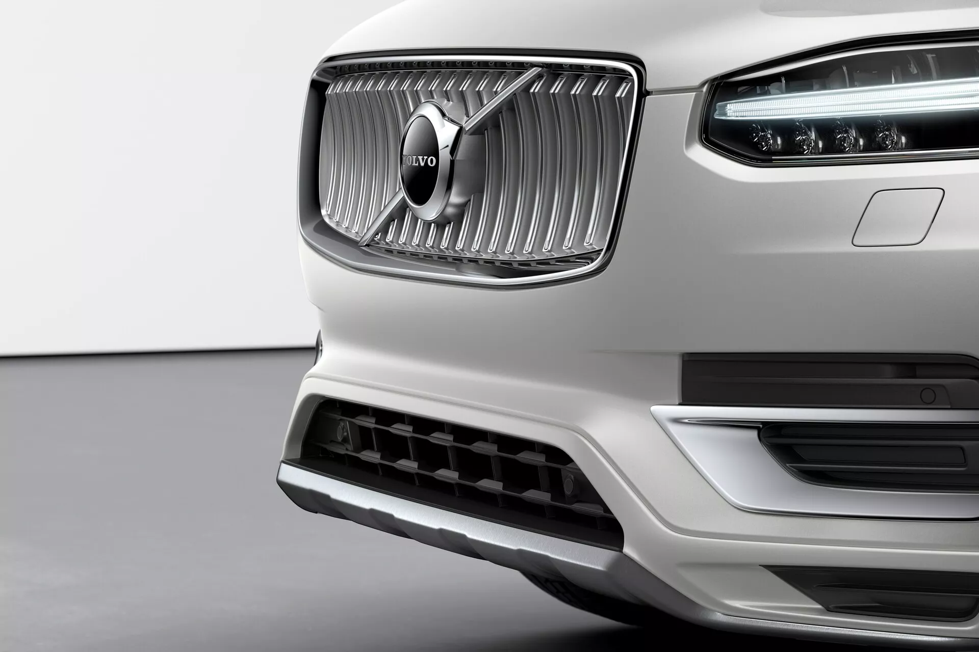 Primer plano de la sección frontal de un Volvo XC90 blanco de segunda generación 2015, destacando su prominente parrilla con rendijas verticales y el distintivo emblema de Volvo en el centro. El acabado cromado de la parrilla contrasta con la textura mate que la rodea. Las luces diurnas LED Martillo de Thor son visibles en el conjunto de faros. El parachoques inferior presenta una gran toma de aire con un borde plateado, lo que sugiere eficiencia aerodinámica y refrigeración del motor. Fotografía tomada desde un ángulo ligeramente bajo, enfatizando la elegancia, calidad de construcción y diseño frontal premium de Volvo.